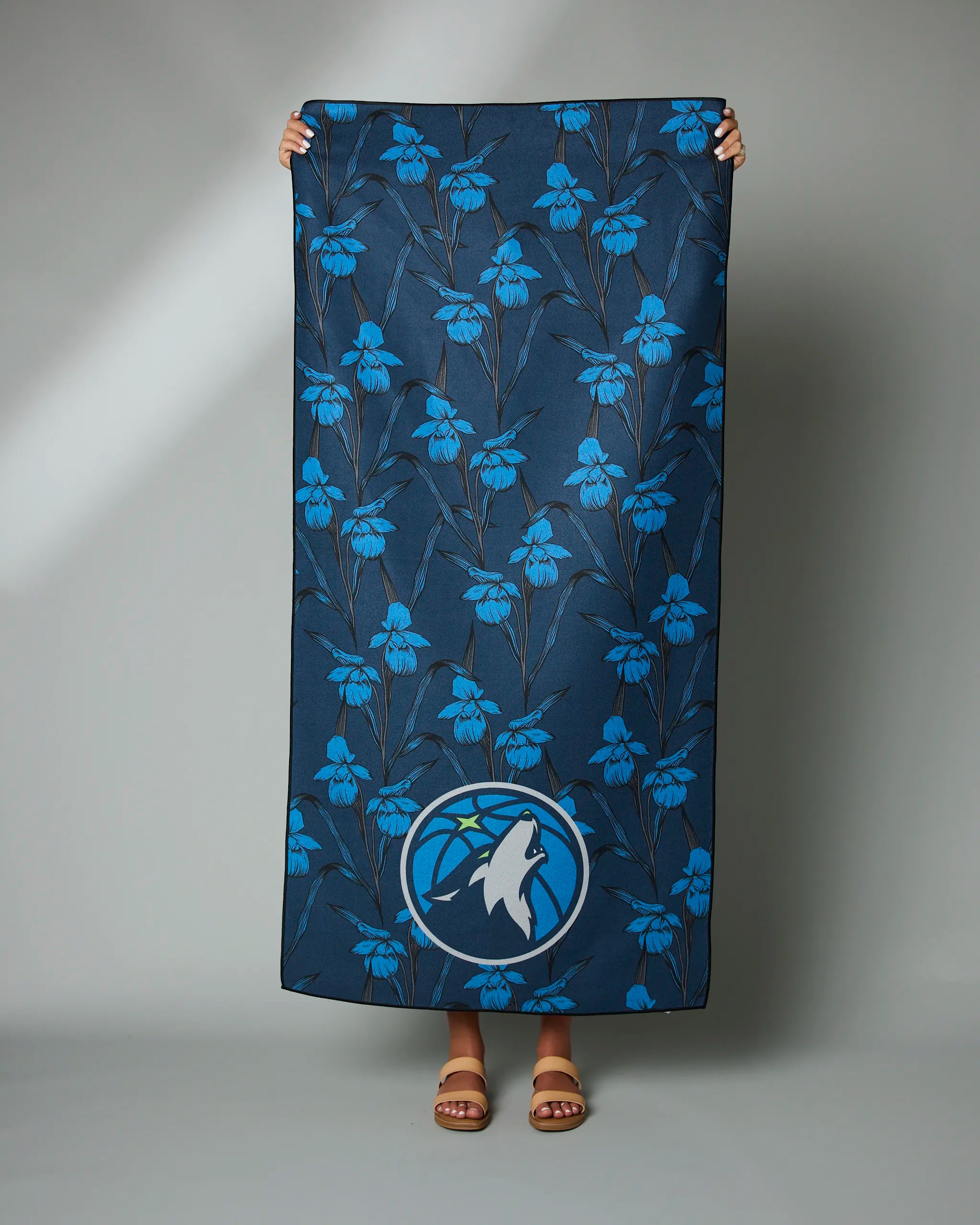 A person displays the Geometry Timberwolves Floral towel, featuring a dark blue base with blue floral patterns and a Timberwolves wolf logo at the bottom. Only their hands and feet in sandals are visible.
