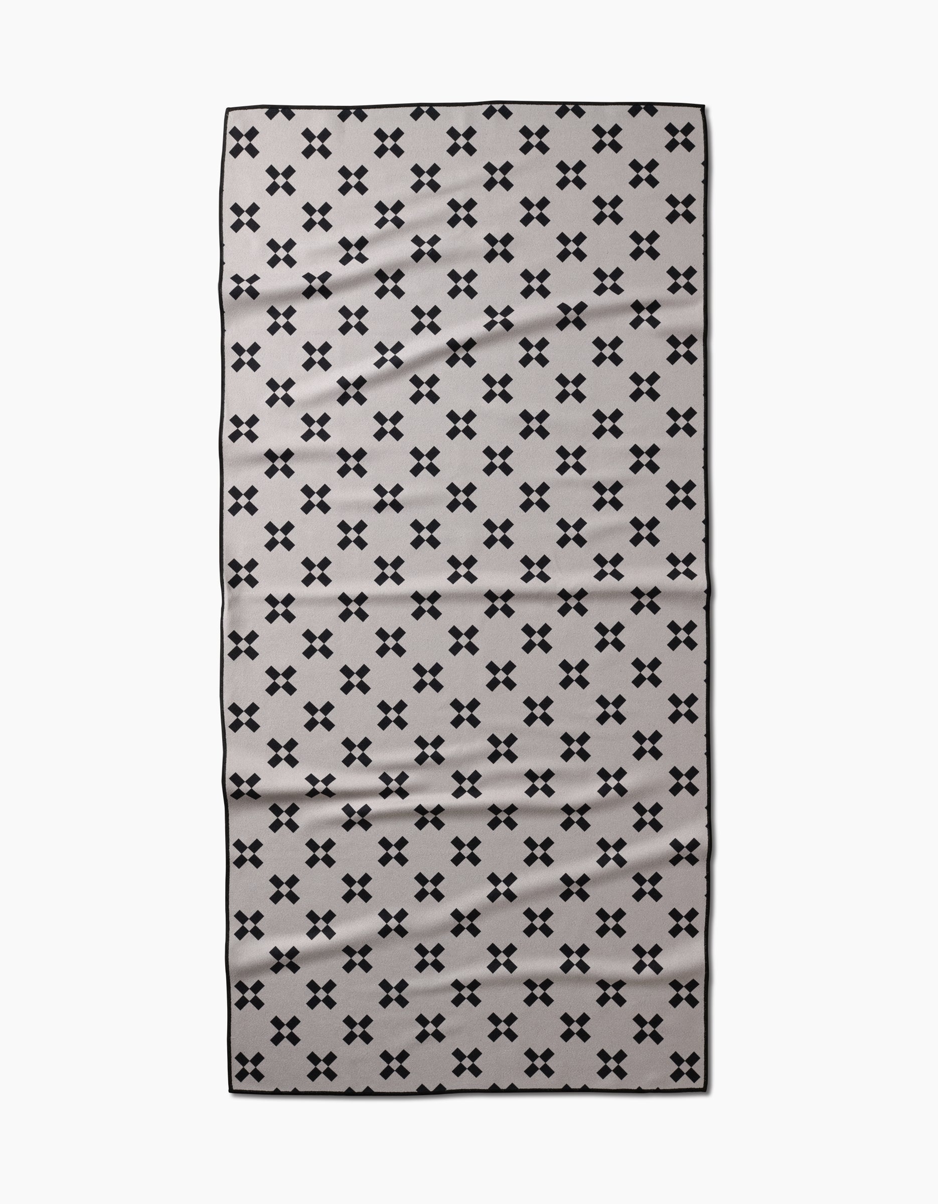 The Tova Beach Towel by Anna Christian is a rectangular, light gray towel with small black X patterns and a thin black border. Made from recycled materials, it is quick-drying and designed for the beach.