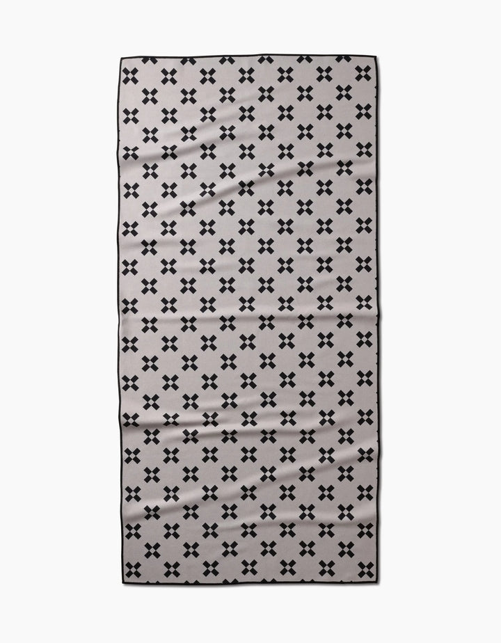 The Tova Beach Towel by Anna Christian is a rectangular, light gray towel with small black X patterns and a thin black border. Made from recycled materials, it is quick-drying and designed for the beach.