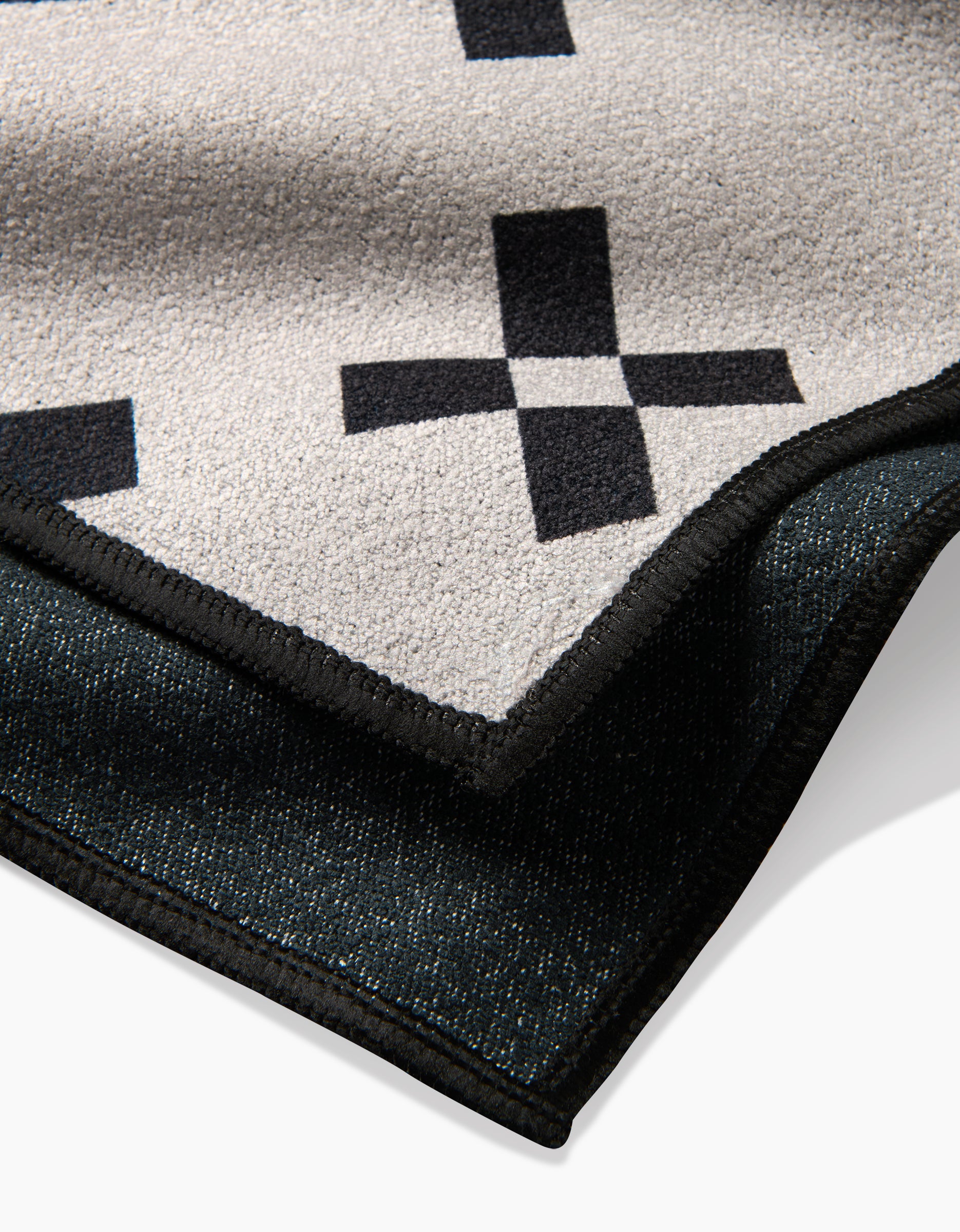 A close-up of the Tova Beach Towel by Anna Christian shows its beige surface with black plus sign patterns, a dark textured reverse, and sleek black trim. Made from recycled materials.