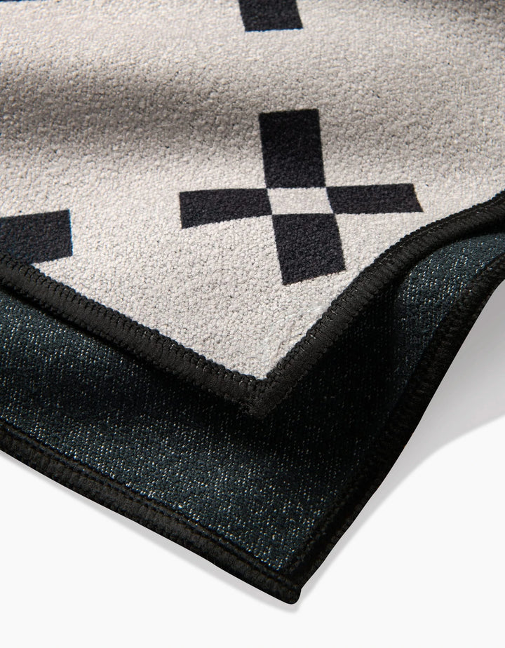 A close-up of the Tova Beach Towel by Anna Christian shows its beige surface with black plus sign patterns, a dark textured reverse, and sleek black trim. Made from recycled materials.