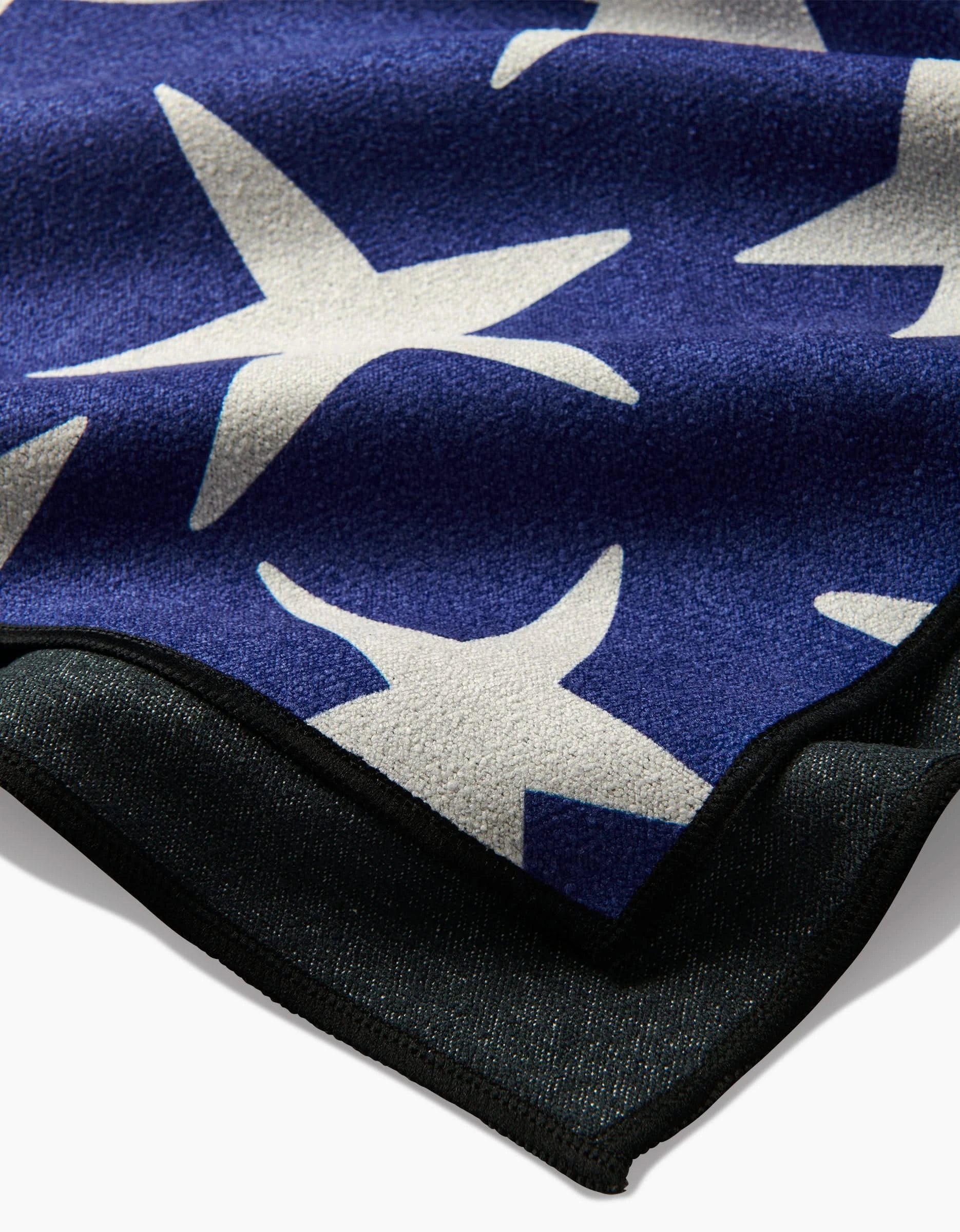 A close-up of the Mindfulnice USA Stars Micro-loop Beach Towel, made from recycled materials, featuring large white five-pointed stars on blue fabric with a black edge trim.