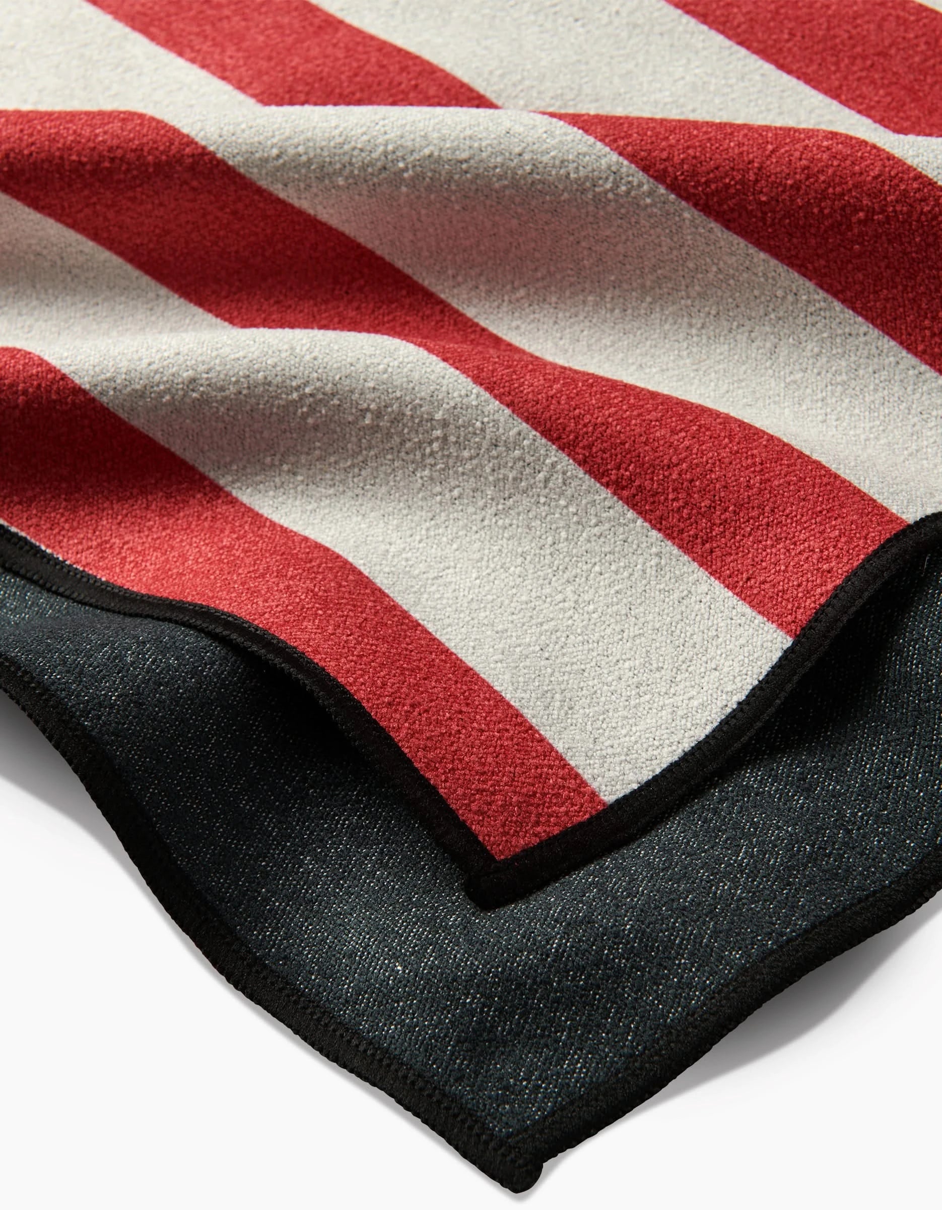 A close-up of a folded Mindfulnice USA Stripes micro-loop beach towel shows bold red and white stripes above dark material with black trim, crafted from quick-drying, recycled materials.