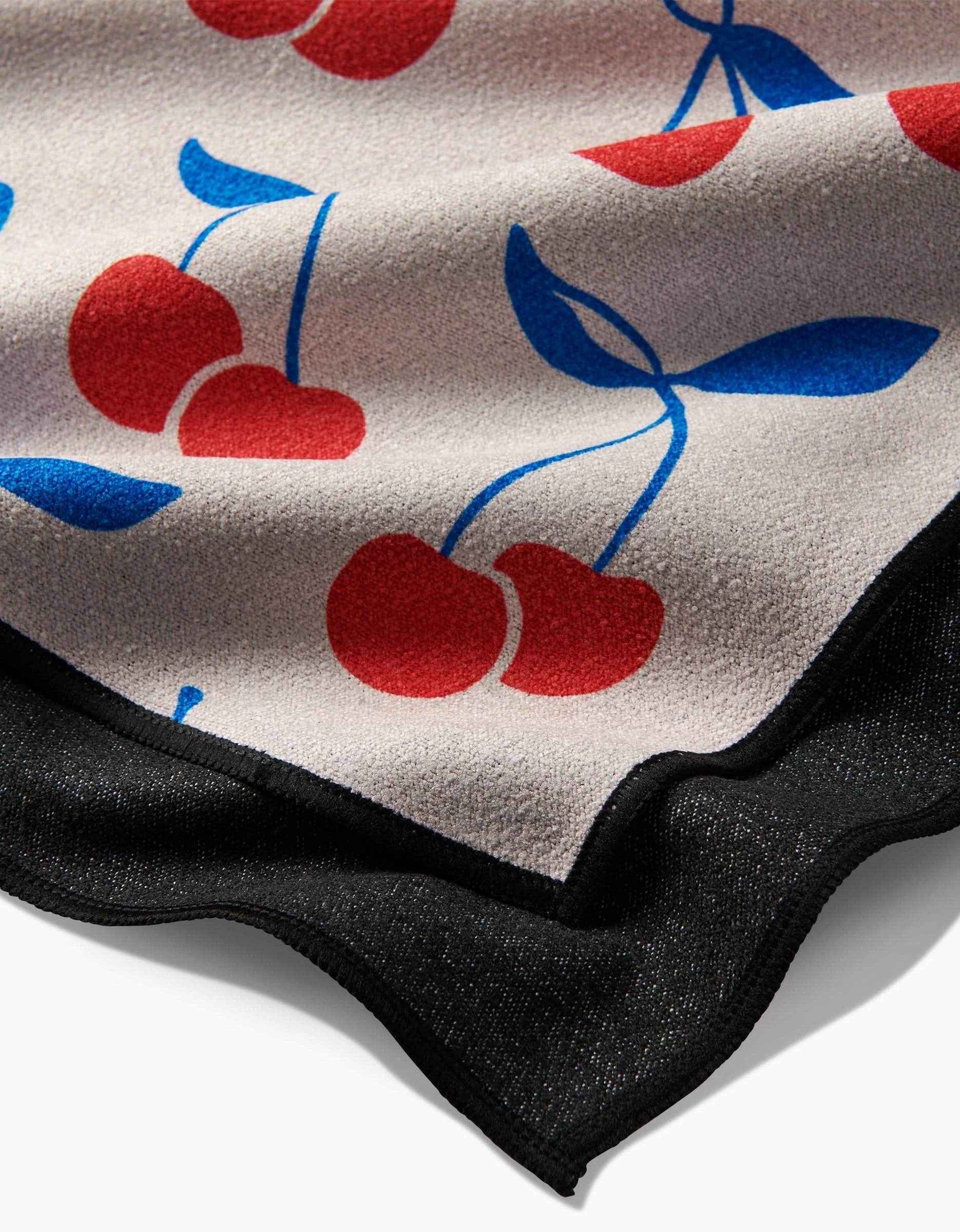 A close-up of Kayla Anns Very Cherry Micro-loop Beach Towel, showing its textured surface, red cherry and blue leaf pattern, and black wavy edge—all crafted from recycled materials.