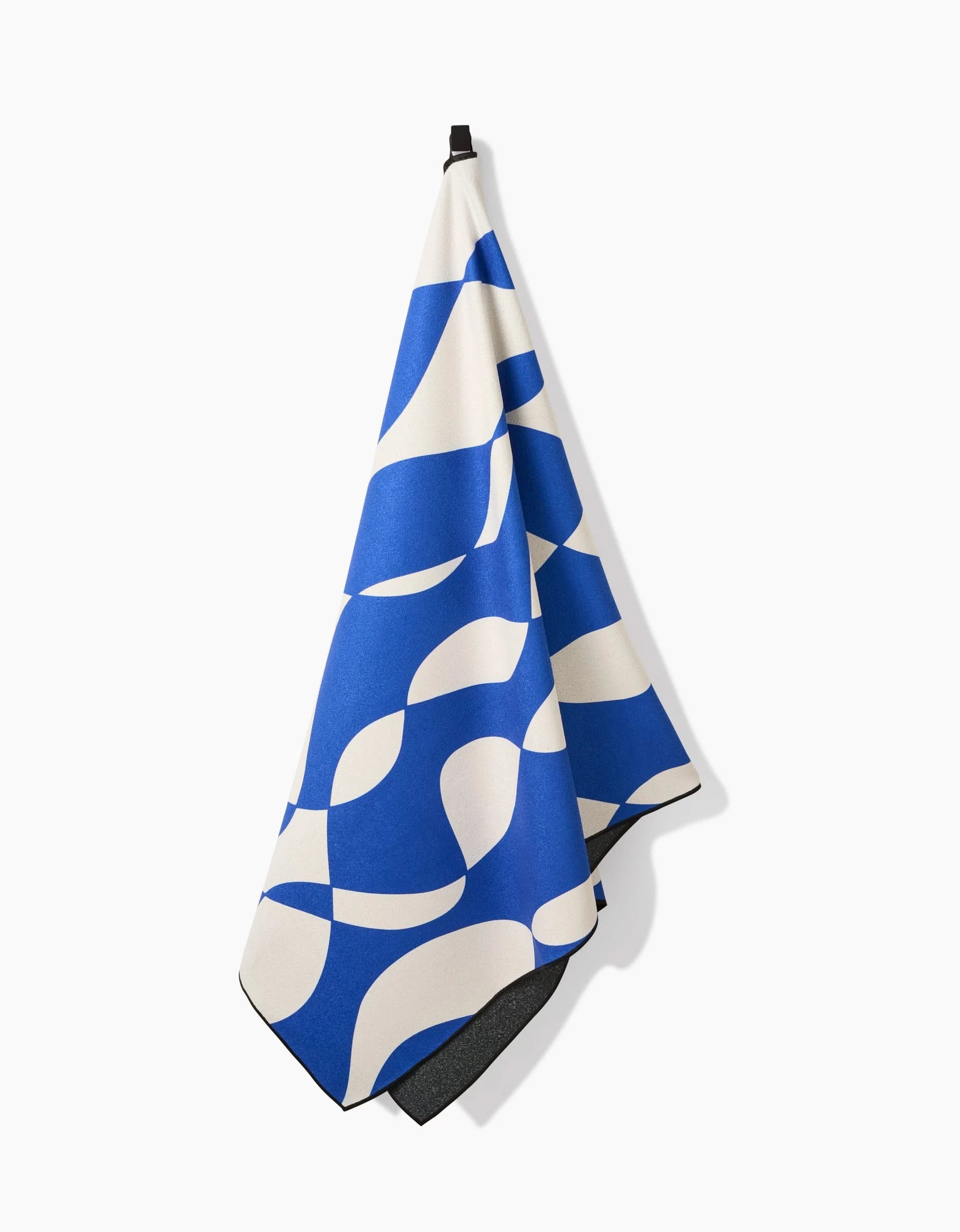 The Wonky Lines micro-loop beach towel by Claire Iglesias, featuring blue and white abstract patterns, hangs from a black hook. Made from recycled materials, this quick-drying towel is both eco-friendly and stylish.