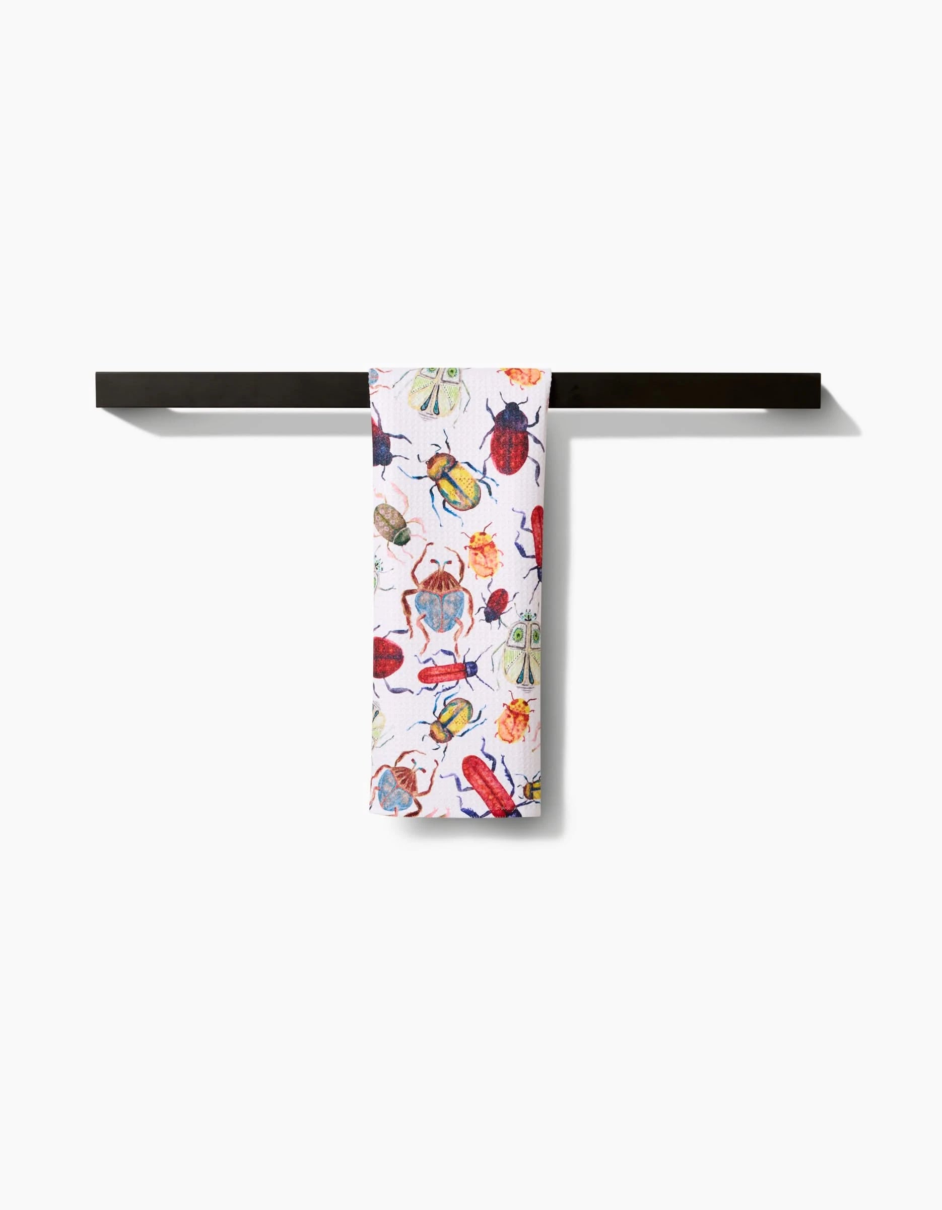 The Taryn Brennan African Beetles white bar towel, featuring colorful illustrated beetles, hangs neatly folded over a simple black towel rack against a plain white background.