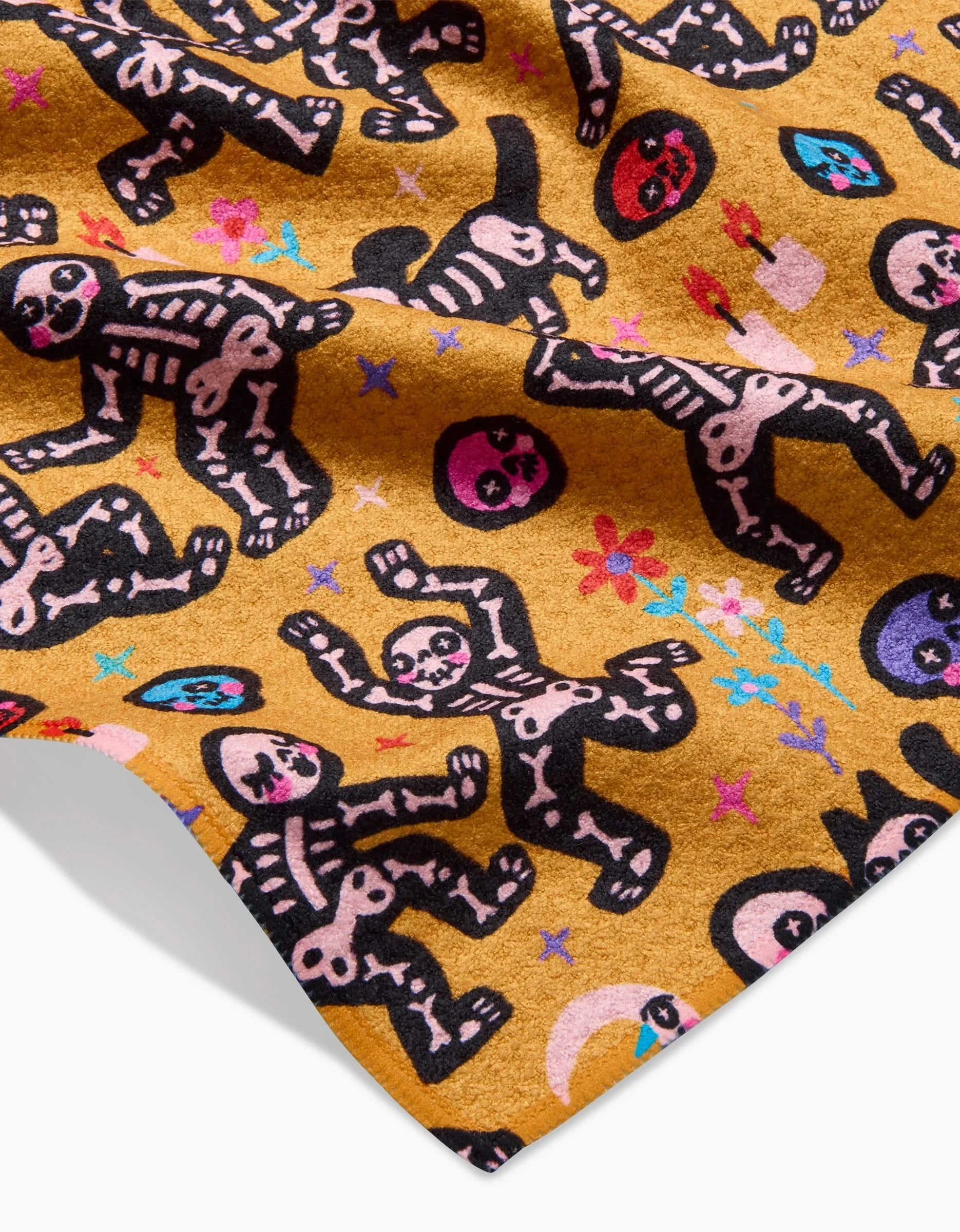 The After Party bar towel by So Lazo is mustard yellow, made from recycled materials, and features playful skeletons, crescent moons, and multicolored flowers in pink, red, blue, and purple outlined in black.