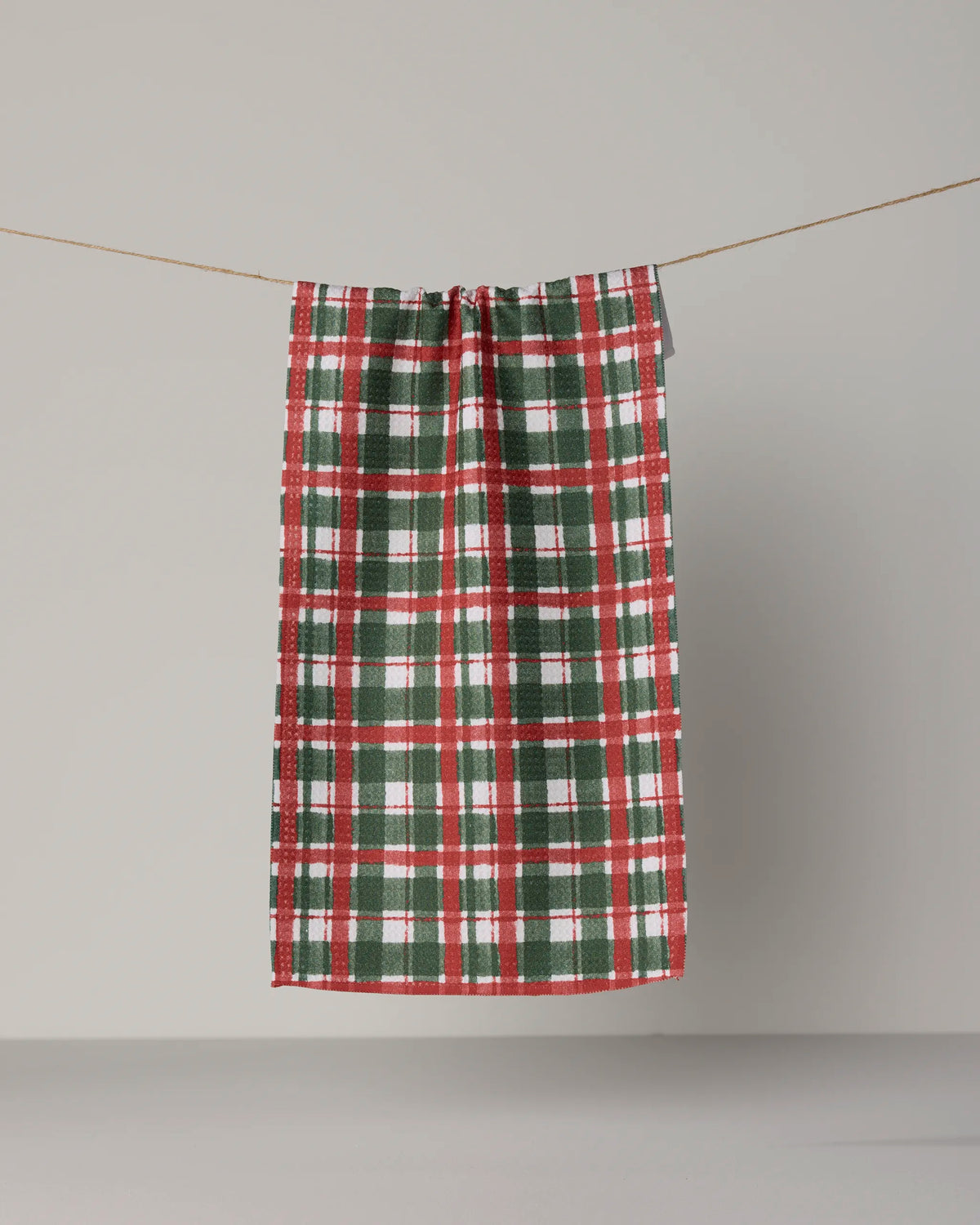 The Alpine Plaid kitchen mini towel by Geometry, featuring red, green, and white stripes in a classic plaid pattern, hangs on a clothesline against a plain, light gray background. Made from recycled materials, this charming accent combines style and sustainability for your kitchen routine.
