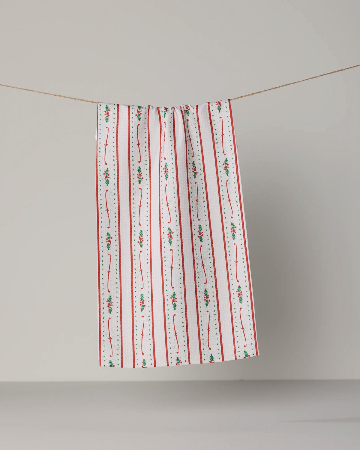 A Geometry “Alpine Trim” mini towel, showcasing red and green floral and stripe patterns on a white background, hangs neatly on a clothesline against a plain, light gray backdrop.