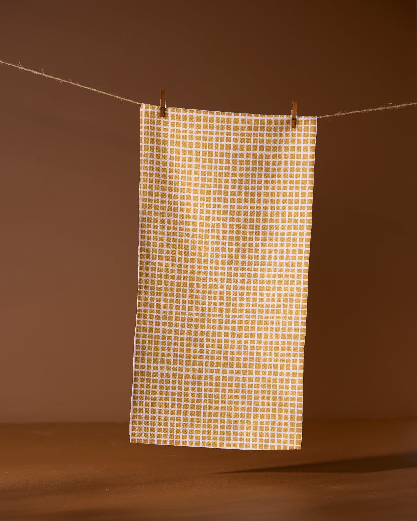 The Geometry Amberline towel, featuring a white and yellow checked design, hangs from a string with two clothespins against a brown background.