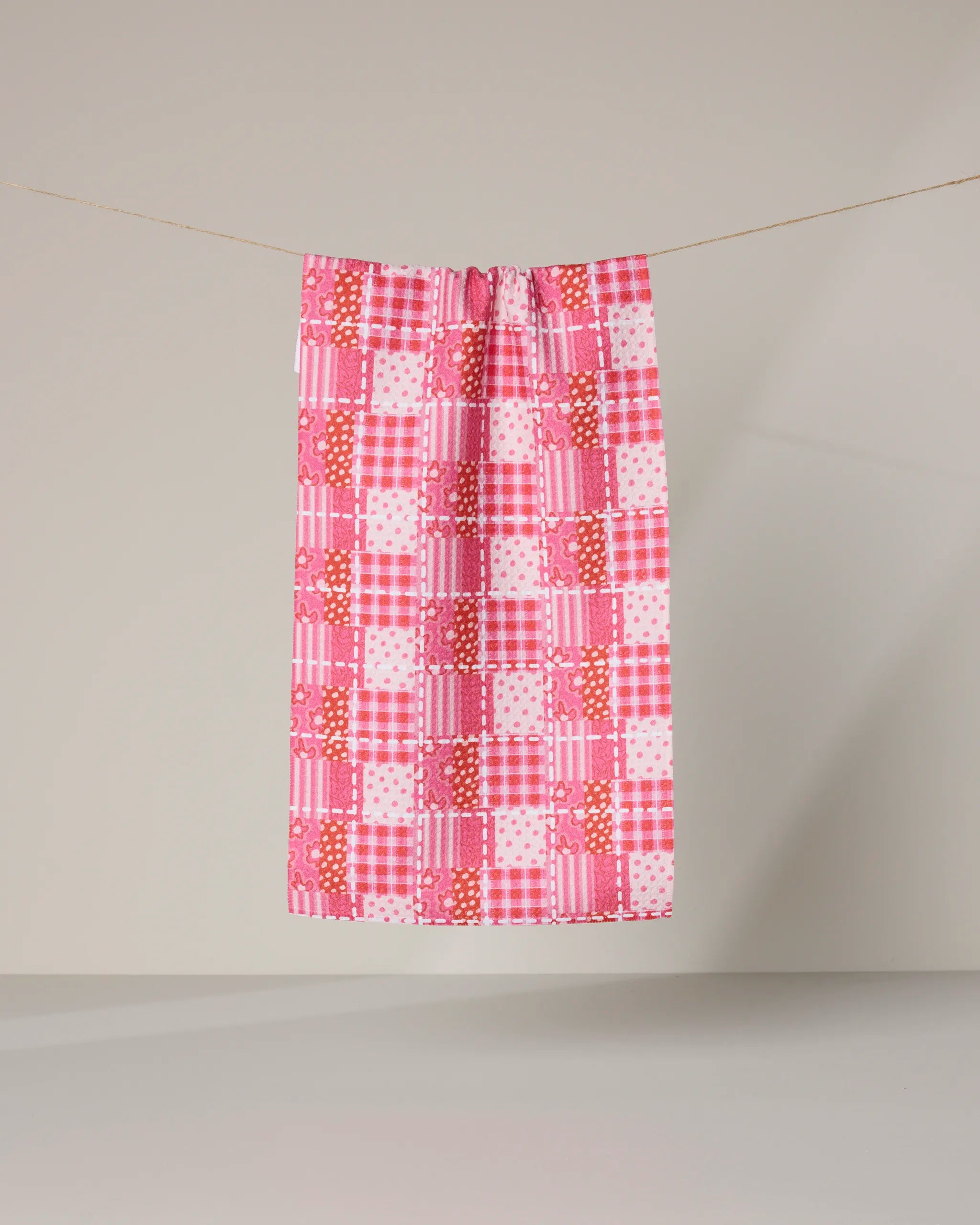 The Geometry Amor Amor bar towel, featuring pink, red, and white checkered patterns, hangs neatly on a clothesline against a light background.