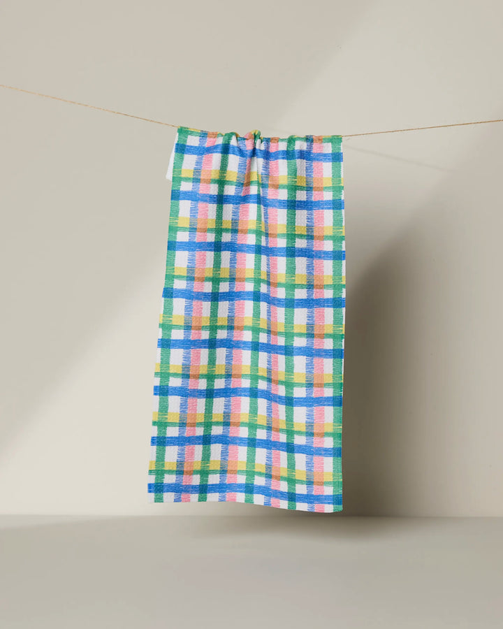 The Geometry Ansel recycled kitchen towel, featuring blue, green, pink, and yellow checkered squares, hangs on a string against a plain light beige background.