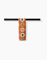 The Megan Roy Apples Acorns necktie, with circular leaf, apple, and grid patterns in brown, yellow, and pink on orange, hangs over a black bar towel.