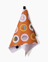 The Megan Roy Apples Acorns recycled kitchen towel features acorns, hearts, and leaves inside pink and yellow circles on orange fabric, shown hanging from a black loop against a white background.