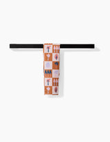 The Autumn Forest recycled kitchen towel by Claire Iglesias, featuring geometric shapes and plant motifs in orange, pink, blue, and white, hangs neatly over a simple black towel bar against a white background.
