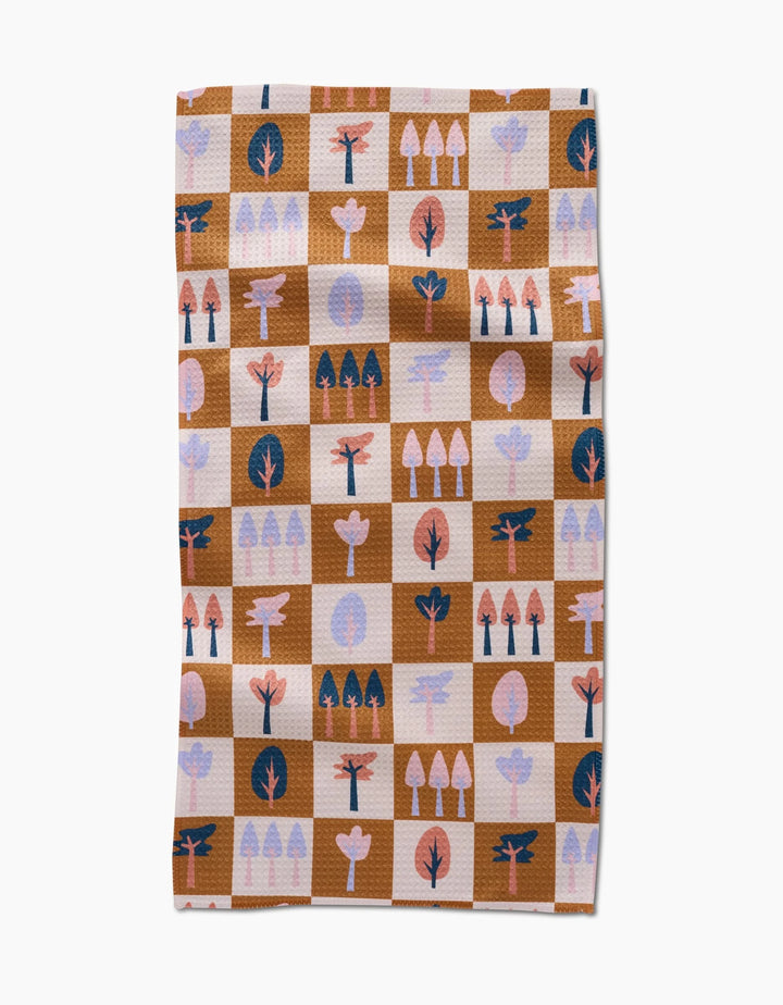 The Autumn Forest kitchen towel by Claire Iglesias is a rectangular recycled towel with a brown and beige checkered pattern, featuring stylized tree illustrations in pink, coral, navy, and light purple on each square.