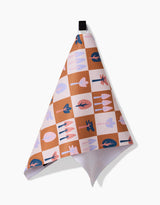 The Autumn Forest recycled kitchen towel by Claire Iglesias features a brown, white, and pink checkered pattern with blue, pink, and red tree and leaf illustrations. Quick-drying and easy to hang from its sturdy black loop.