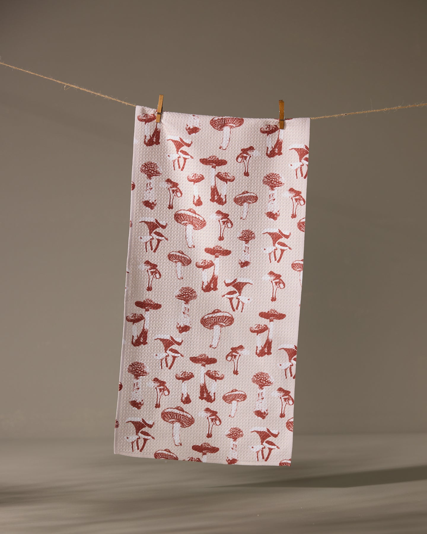 The Geometry Autumn Mushrooms towel, featuring a red mushroom pattern on white, hangs from a clothesline with two wooden clothespins against a neutral background.