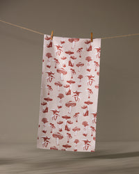 The Geometry Autumn Mushrooms towel, featuring a red mushroom pattern on white, hangs from a clothesline with two wooden clothespins against a neutral background.