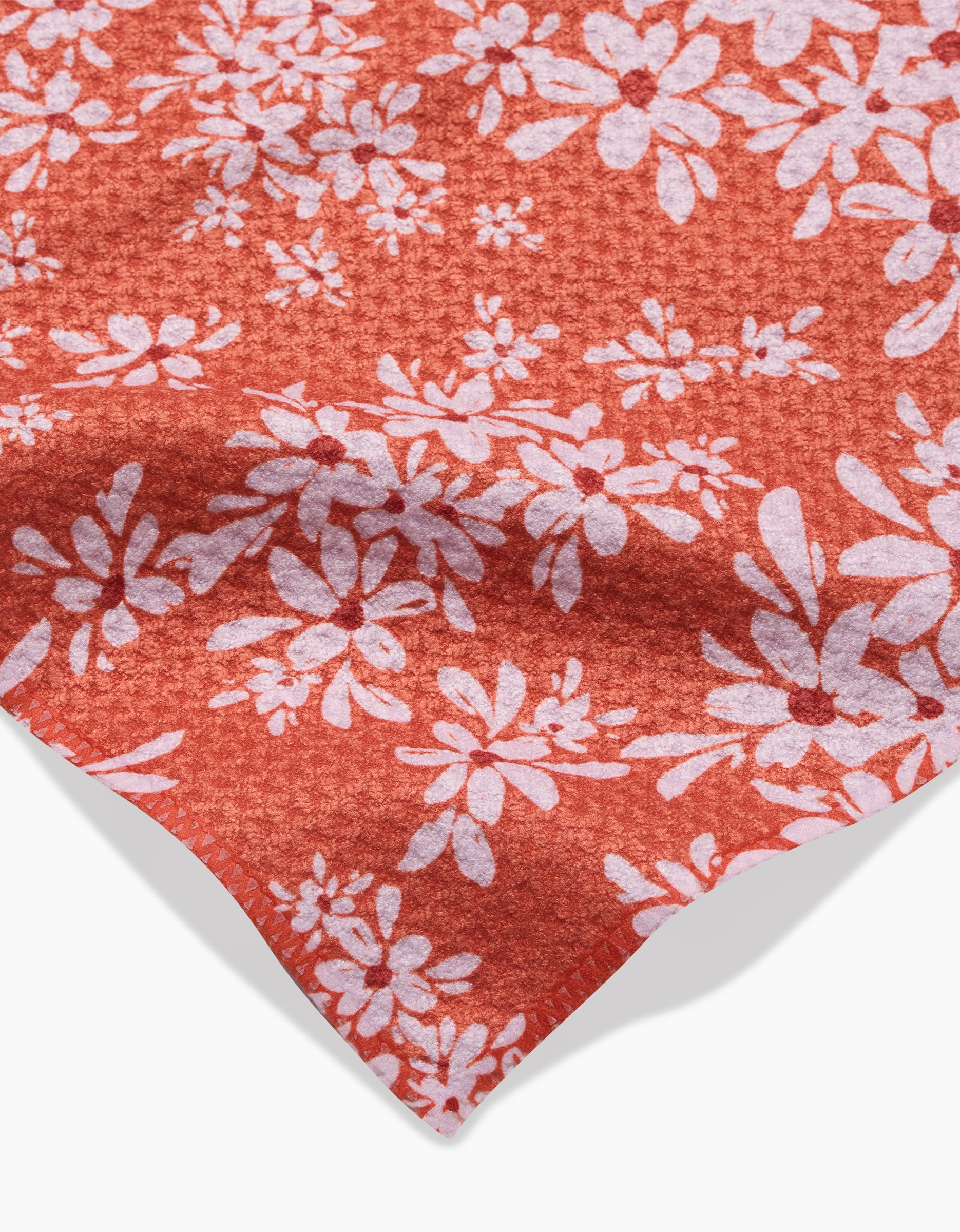 A close-up of the Ashley Celena Autumn Reverie recycled kitchen towel, showing its red fabric with a white daisy-like floral pattern and textured finish, gently folded at the corner.