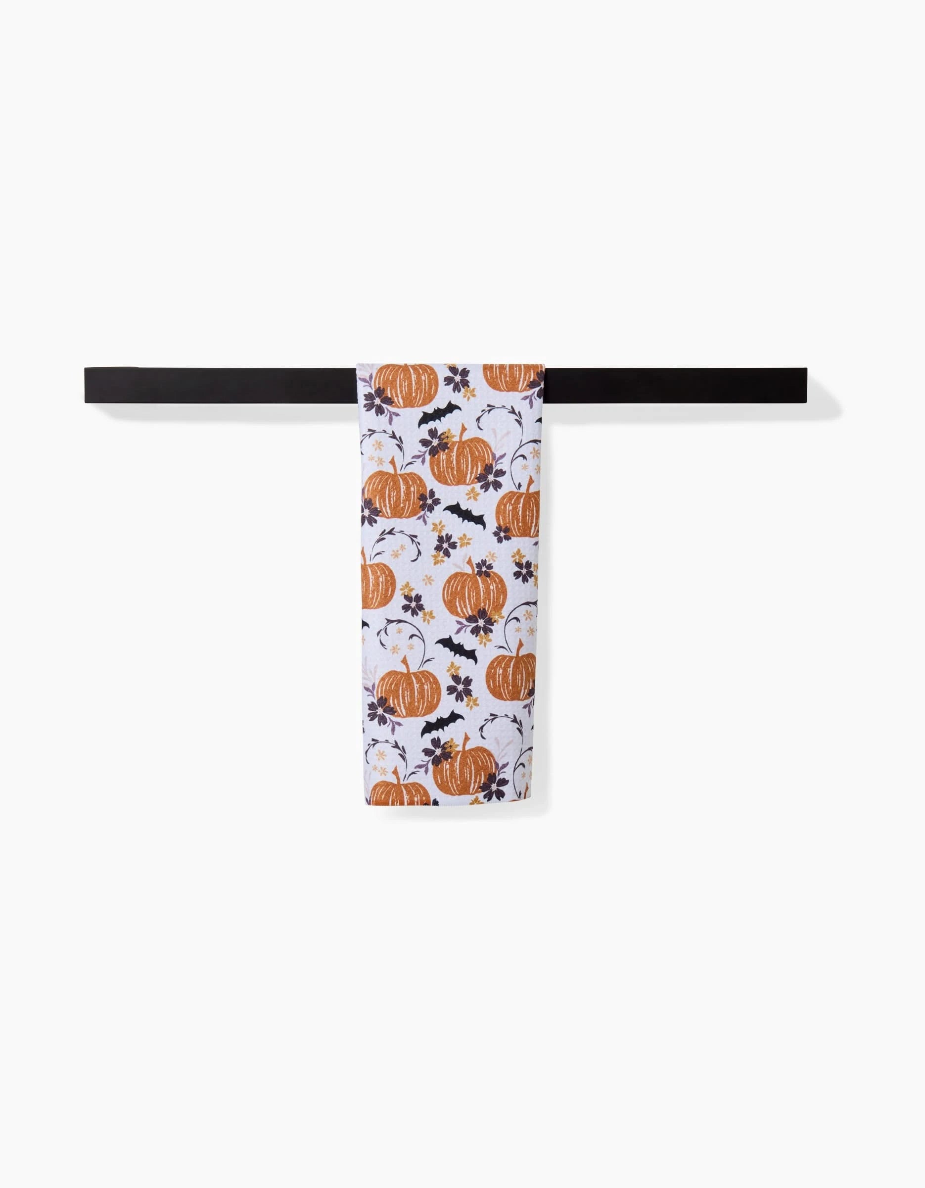 A white ultra-absorbent Bats In The Pumpkin Patch towel by Ashley Celena, featuring an orange pumpkin and leaf print, hangs neatly on a dark horizontal towel bar against a plain white background.