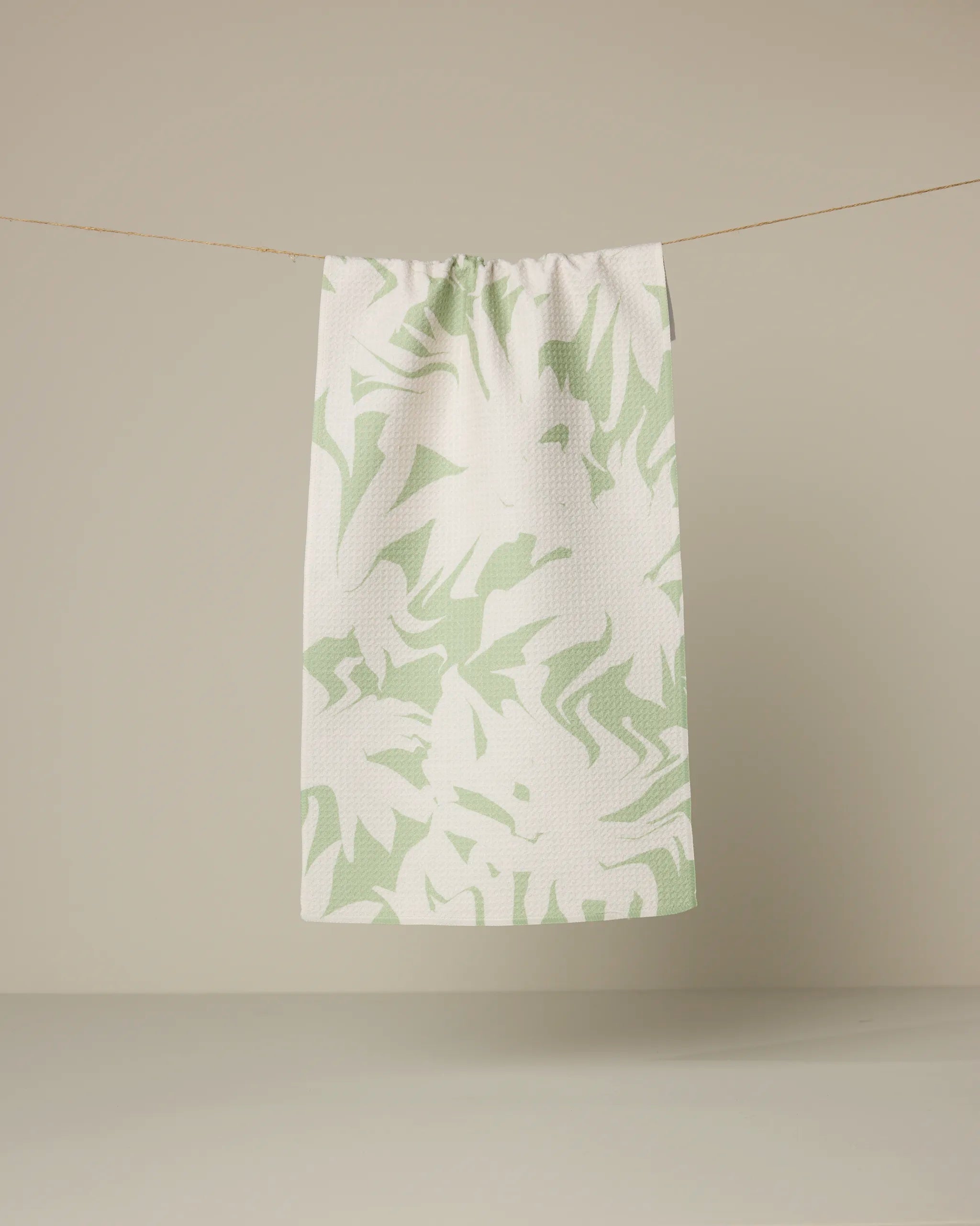 The Geometry Baysage towel, featuring a light green abstract leaf pattern on white, hangs from a string against a plain beige background.