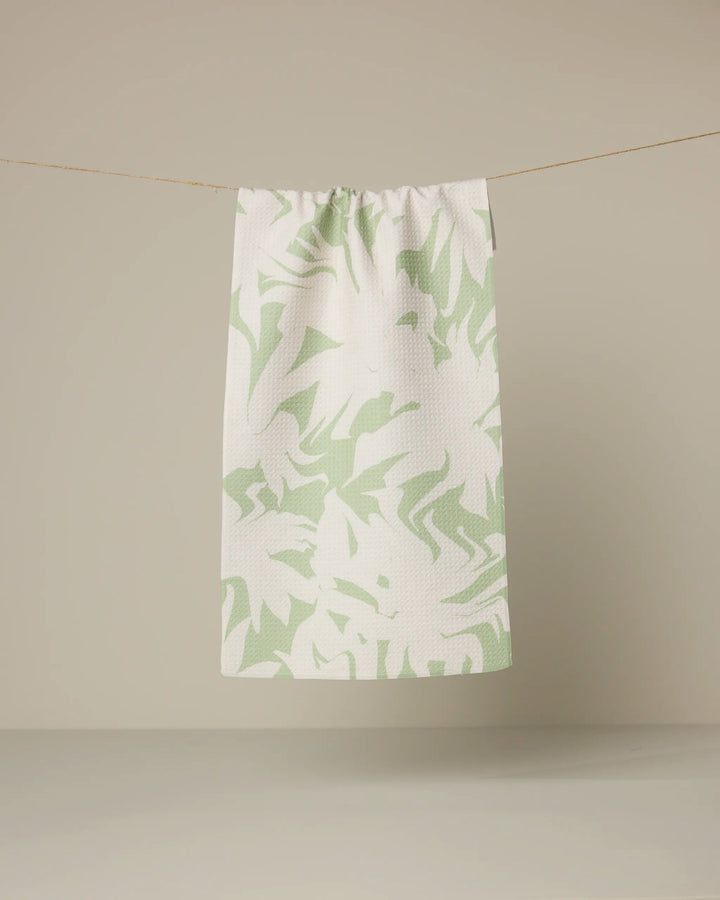 The Geometry Baysage towel, featuring a light green abstract leaf pattern on white, hangs from a string against a plain beige background.