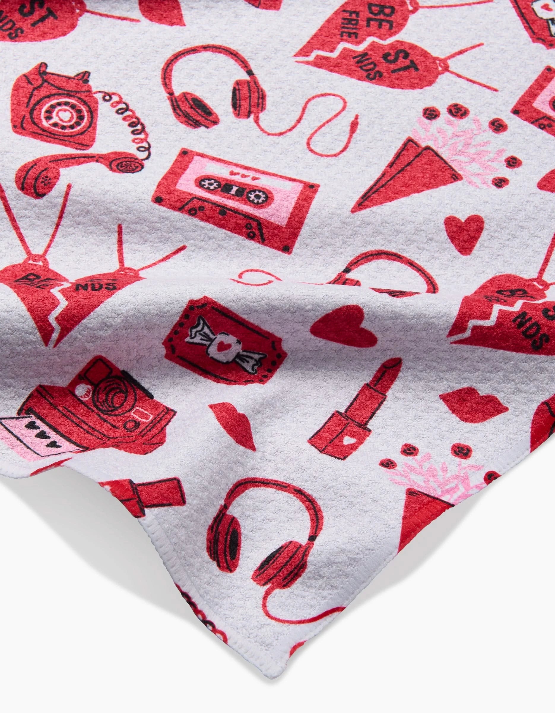 The Best Friends kitchen towel by Charlie Rowan Designs is white and made from recycled materials, featuring playful red and pink retro illustrations like phones, cassette tapes, lipstick, headphones, cameras, candy, hearts, and envelopes.