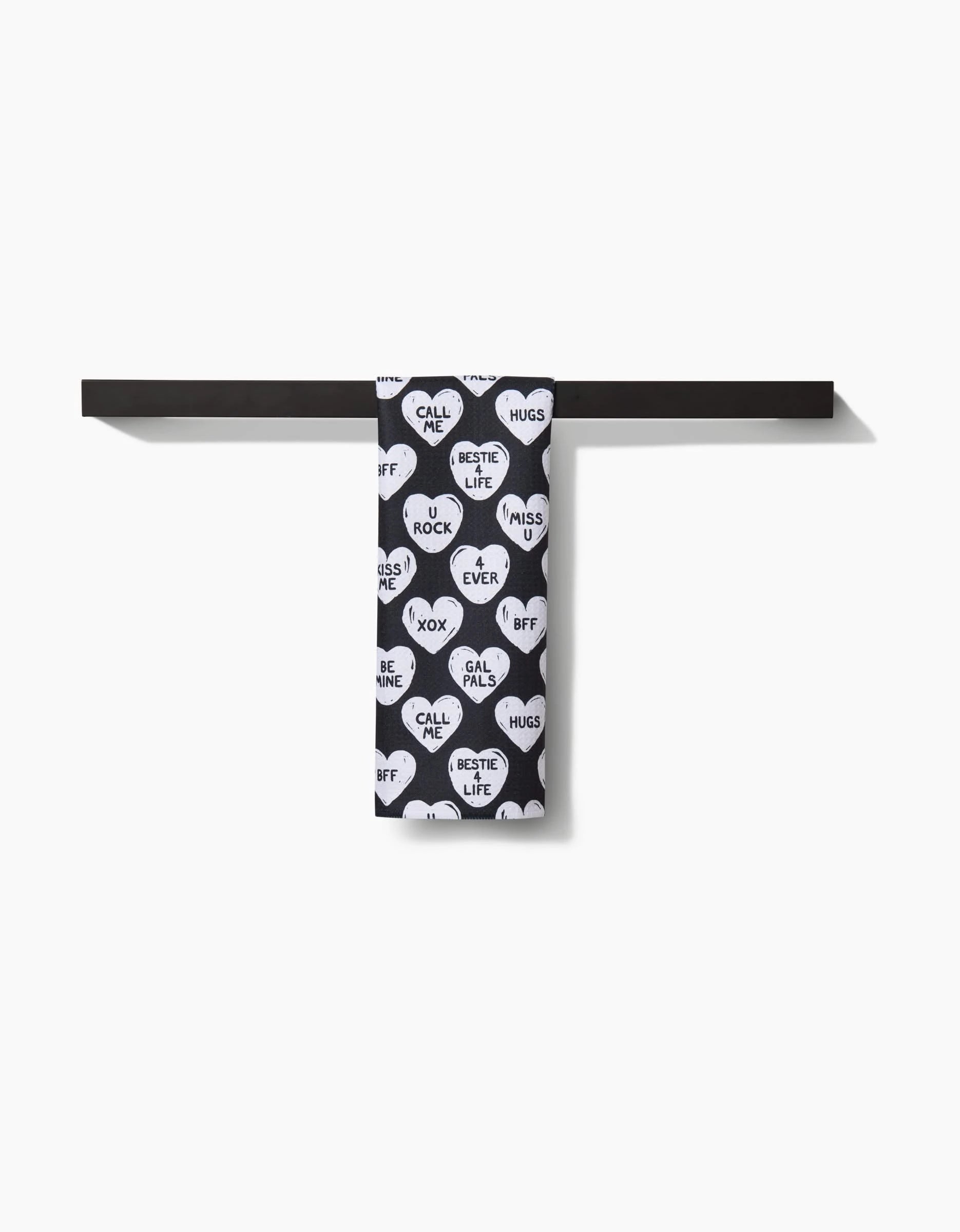 The Hufton Studio BFF Candy Hearts bar towel, black with white heart shapes and playful messages like BFF, HUGS, and CALL ME, hangs neatly on a dark bar against a plain white background.