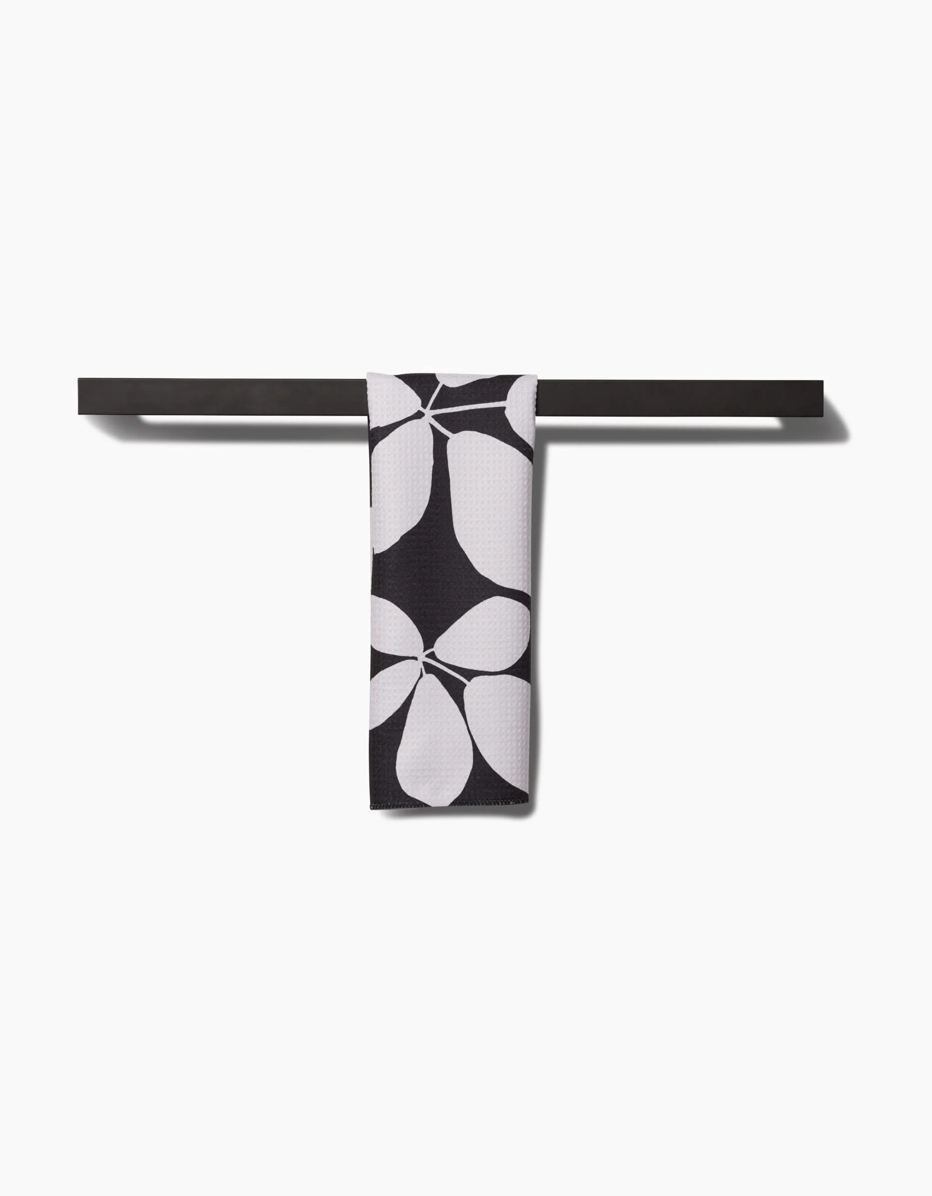 A black Blackberry bar towel by PrintedPlant, made from recycled materials, hangs centered on a black bar and shows a bold white petal or leaf pattern, all set against a plain white background.