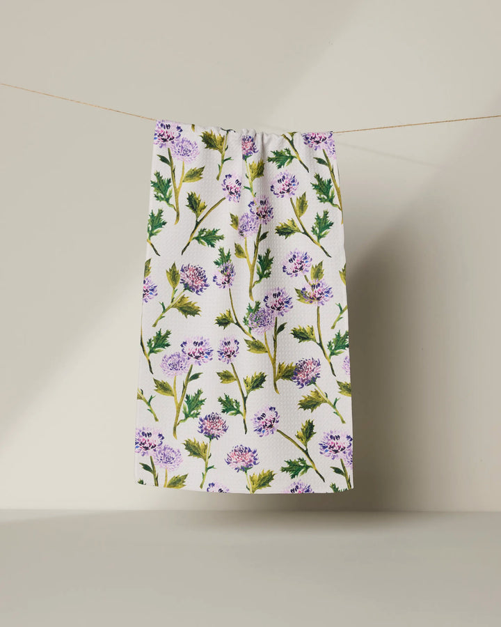 The Geometry "Blooming Thistle" towel, featuring a white, ultra-absorbent fabric with purple flower and green leaf patterns, hangs from a string against a plain light background.