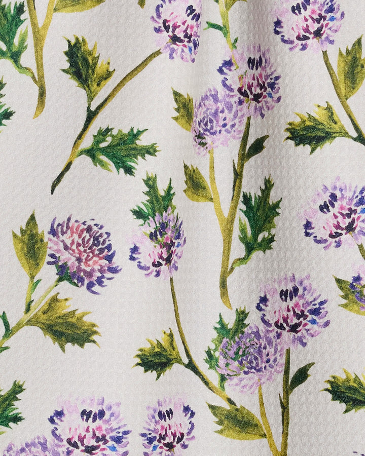 A close-up of Geometry's Blooming Thistle fabric—a textured white material made from recycled fibers, featuring watercolor-style purple thistles and green leaves on yellow stems—ideal for an ultra-absorbent or stylish bar towel.