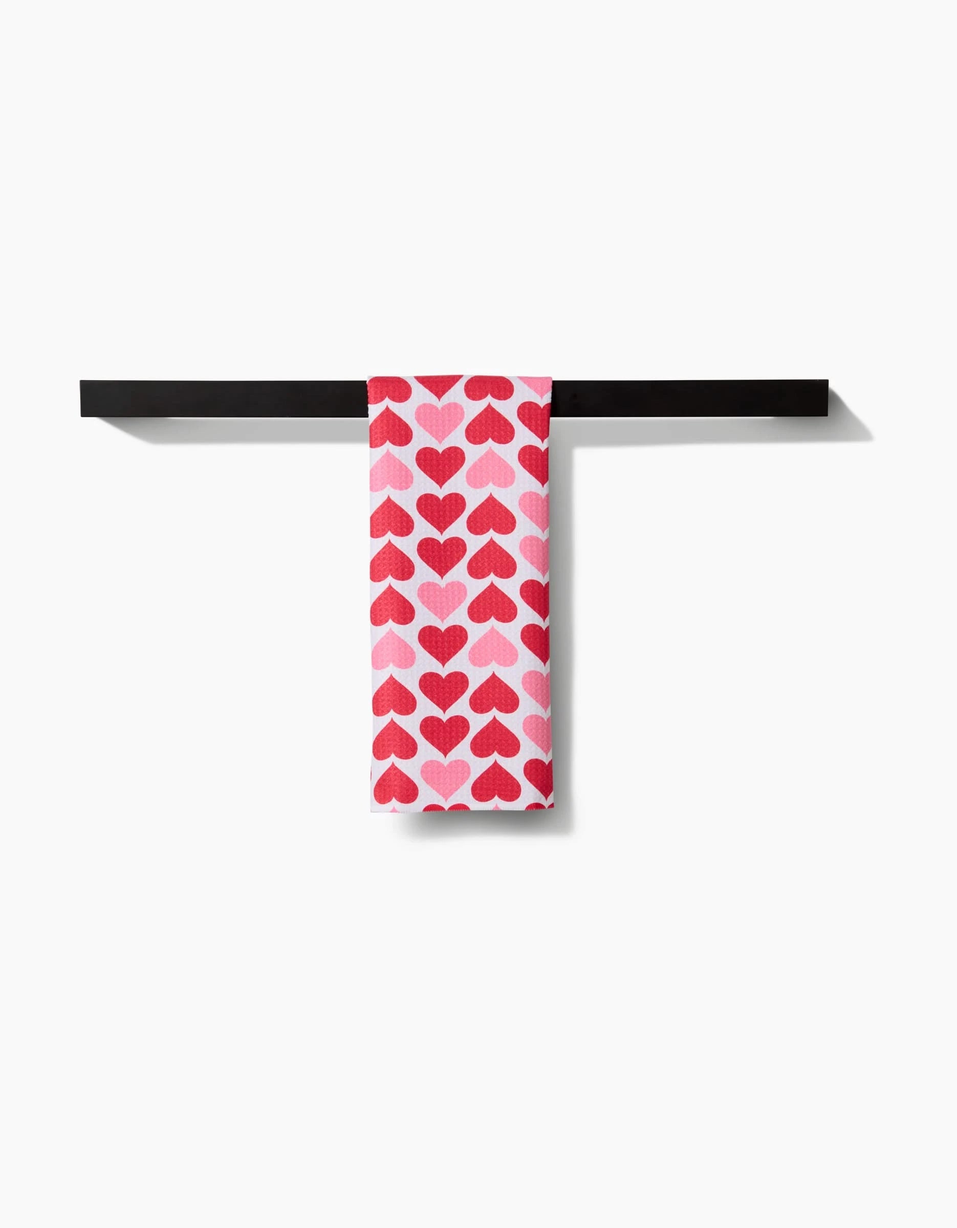 The Blushing Hearts kitchen towel by Danelys Sidron, ultra-absorbent and white with red and pink heart patterns, is neatly folded over a black towel bar against a white background.