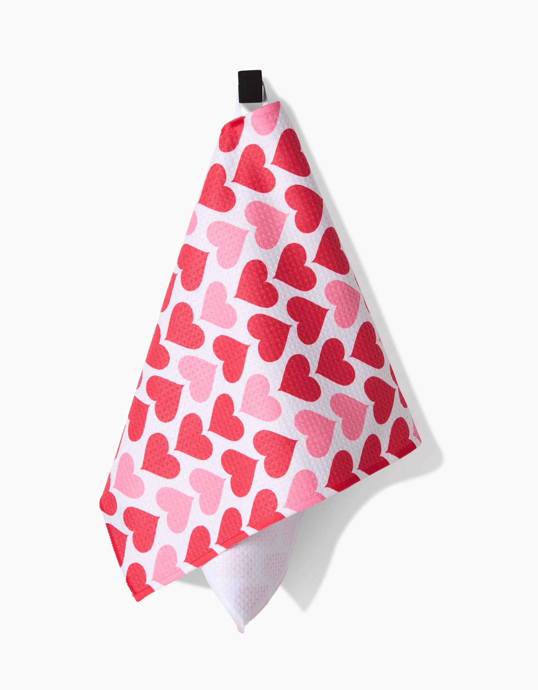Blushing Hearts by Danelys Sidron is an ultra-absorbent pink bar towel featuring overlapping red and pink hearts, hung from a black loop. Made from recycled materials, it blends style and sustainability for your kitchen.