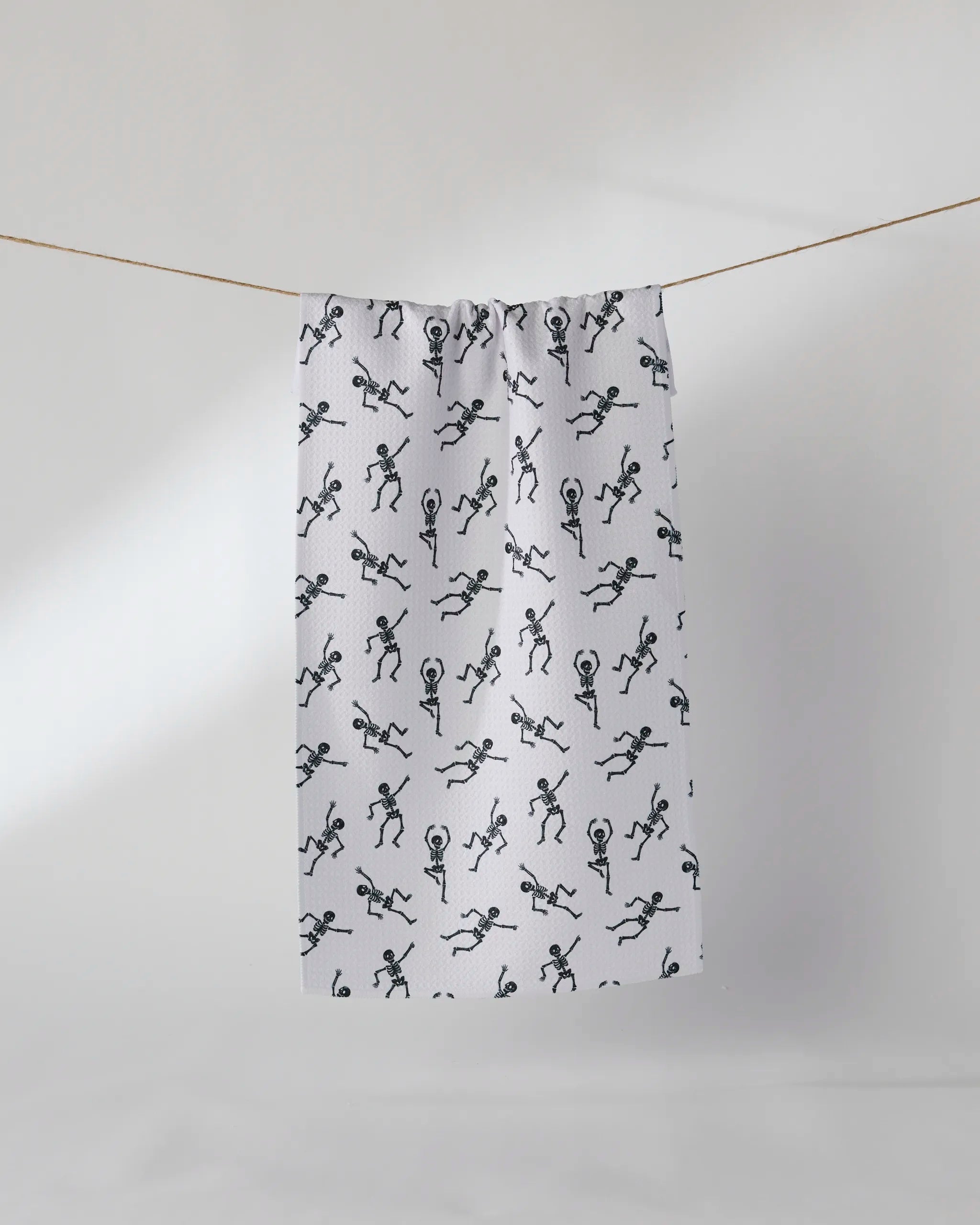 The Geometry Bone Jamboree White towel, made from recycled materials and patterned with playful black stick figures, hangs on a clothesline against a plain, light background.