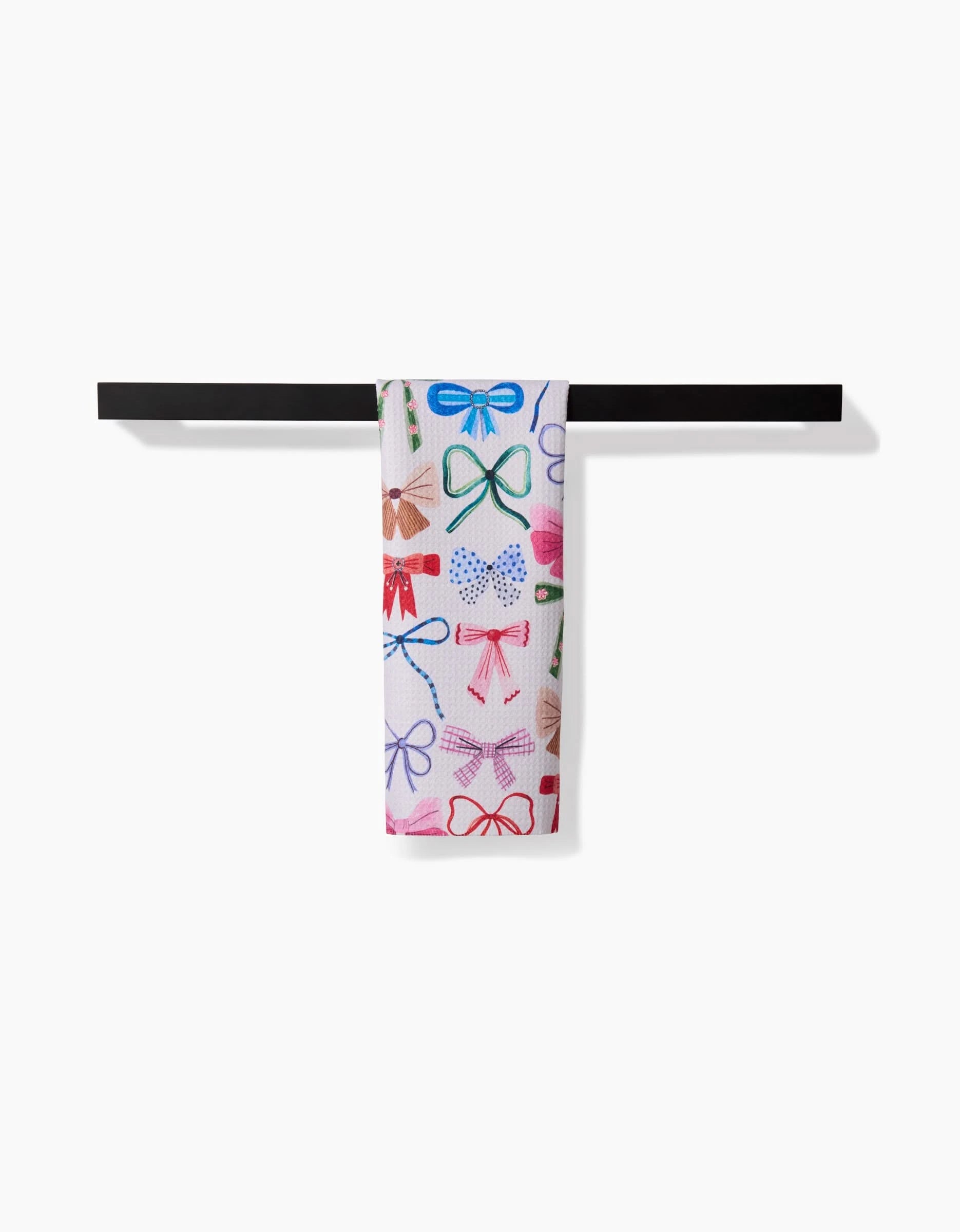 The Daria Solak Bows towel, featuring ultra-absorbent white fabric and vibrant multicolored bow designs, hangs neatly over a black horizontal rod against a plain white background.
