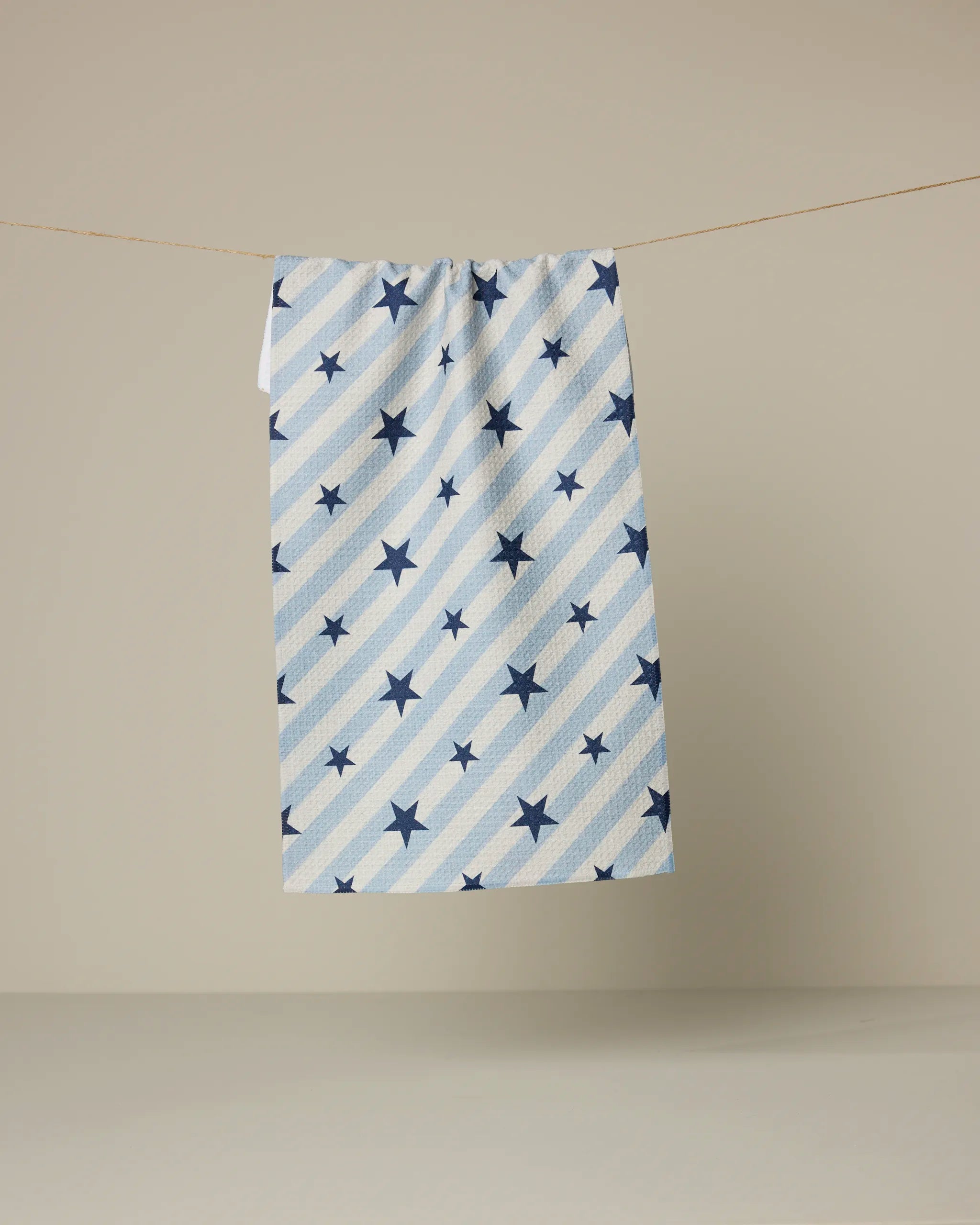 The Geometry Bright Stars towel, featuring blue and white diagonal stripes with blue stars, hangs on a clothesline against a plain beige background.
