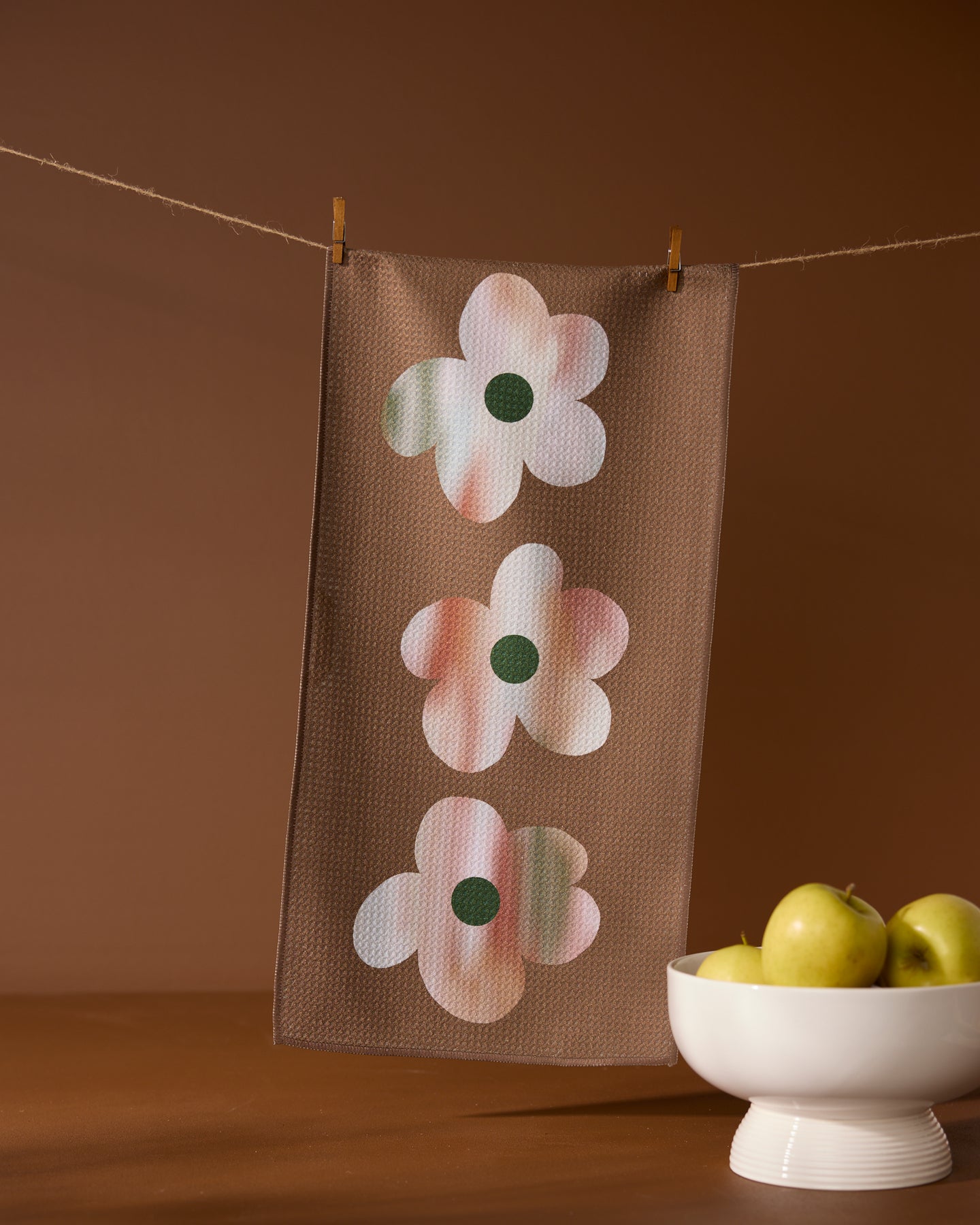 The Brushed Bouquet Brown towel by Geometry, featuring three abstract pastel flower designs, hangs from a clothesline with clothespins. Below it, a white bowl of green apples rests on a surface against a warm brown backdrop.