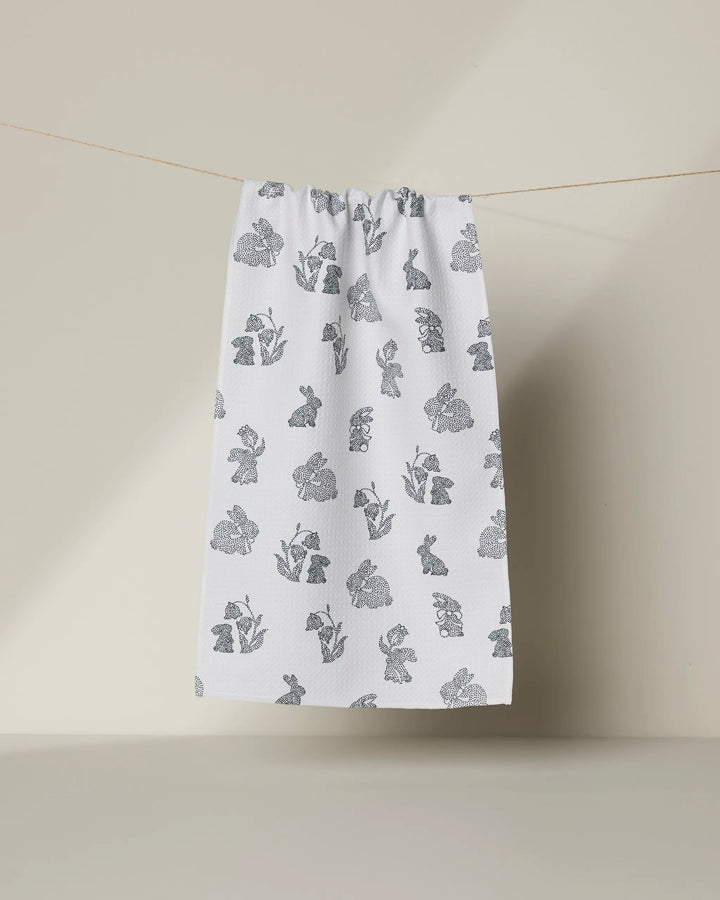 The Geometry "Bunny Dots" white recycled kitchen towel, featuring gray floral and bunny prints, hangs from a string against a plain light beige background.