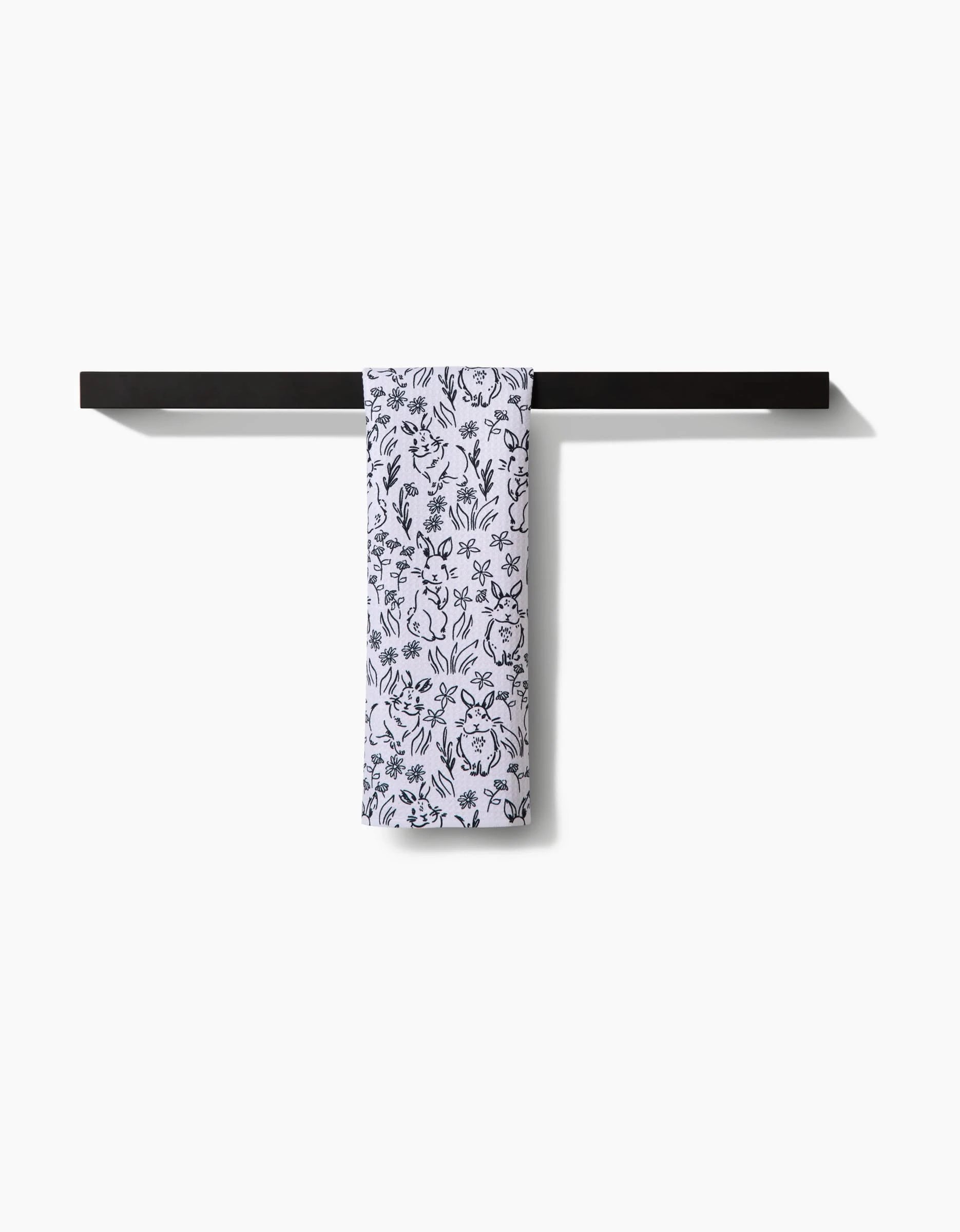 The Bunny Rabbit Garden towel by Charlie Rowan Designs, featuring a black floral pattern, hangs neatly folded over a sleek black towel bar against a plain white background.