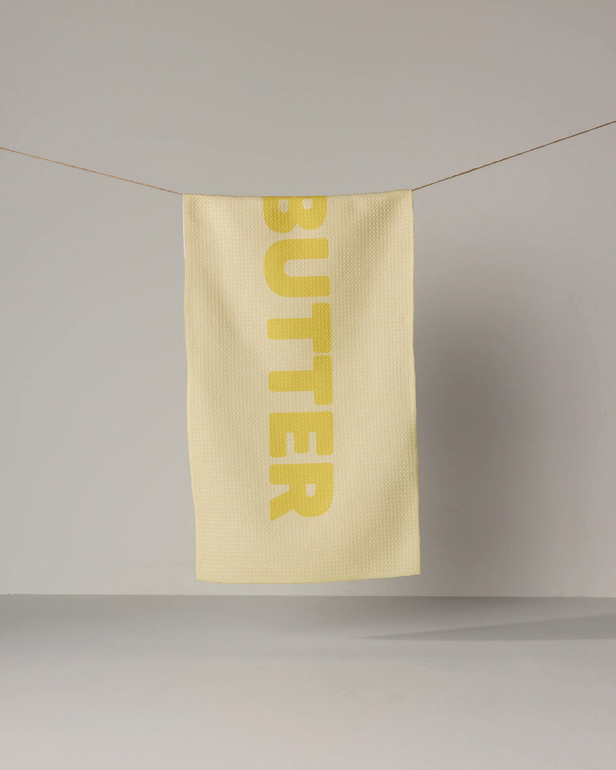 A light yellow GEOMETRY Butter kitchen towel in Geoweave fabric hangs on a string with the word BUTTER printed vertically in large, dark yellow letters.