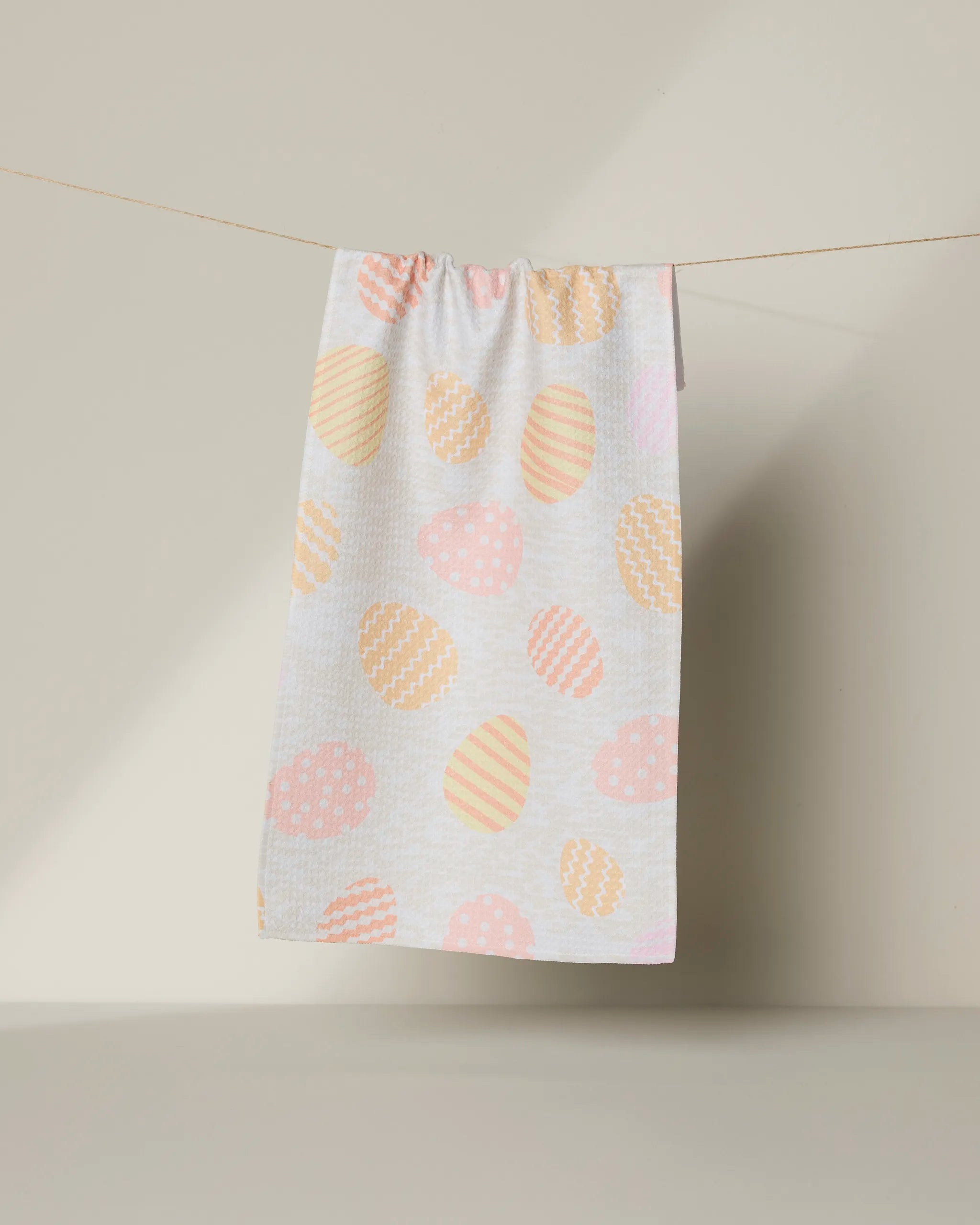 The Geometry Buttercream Easter bar towel, made from recycled materials, showcases pastel-patterned Easter eggs hanging on a clothesline against a light beige background.