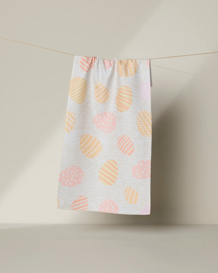 The Geometry Buttercream Easter bar towel, made from recycled materials, showcases pastel-patterned Easter eggs hanging on a clothesline against a light beige background.