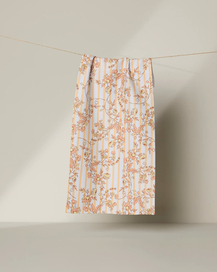 The Geometry “Buttercup Vine” bar towel, made from recycled materials and featuring white fabric with vertical blue stripes and an orange floral pattern, hangs on a string against a plain beige background.