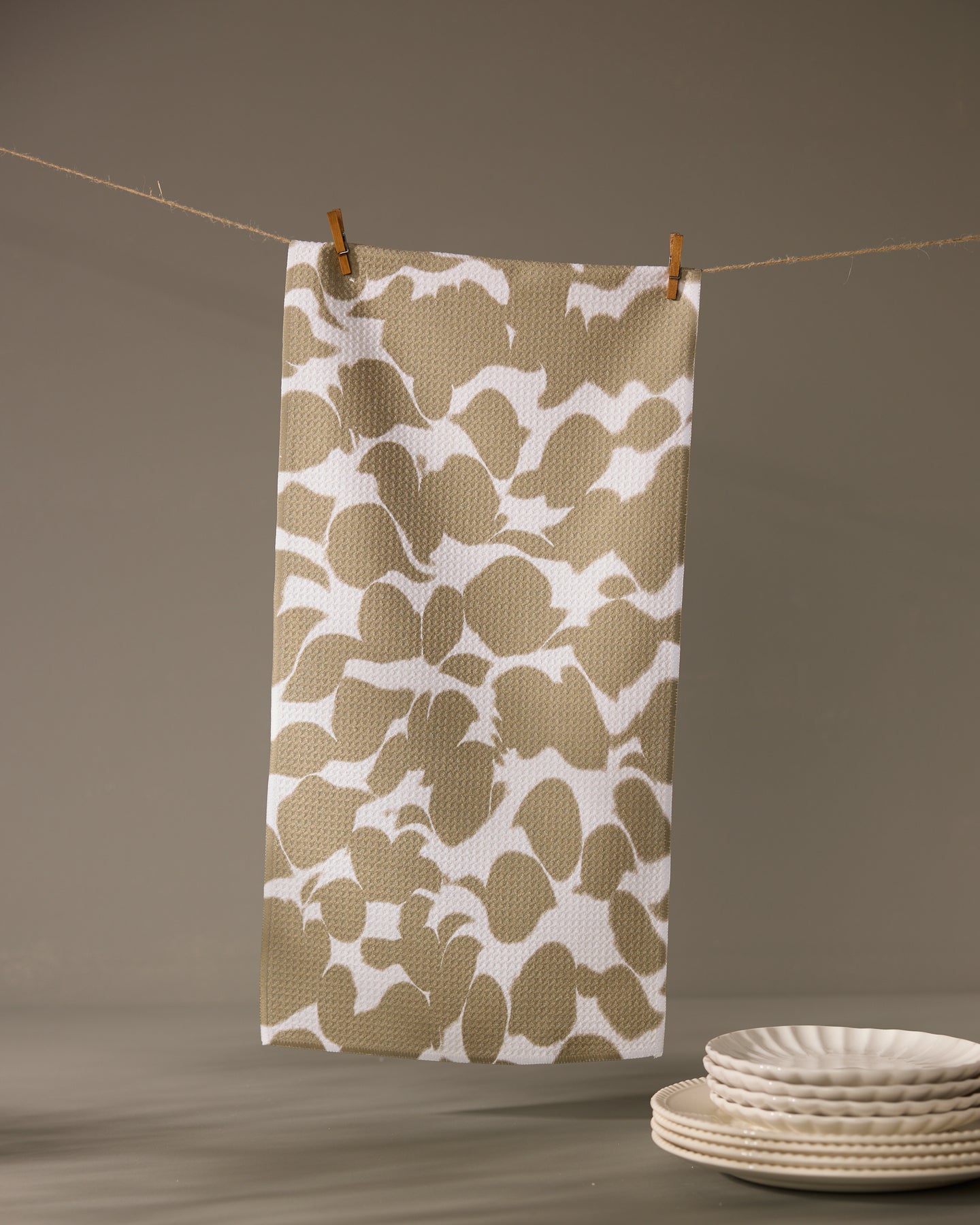 The Geometry Canopy bar towel, featuring a beige and white pattern and ultra-absorbent design, hangs from a string with clothespins. In the foreground, white ceramic plates are stacked neatly on a gray surface.