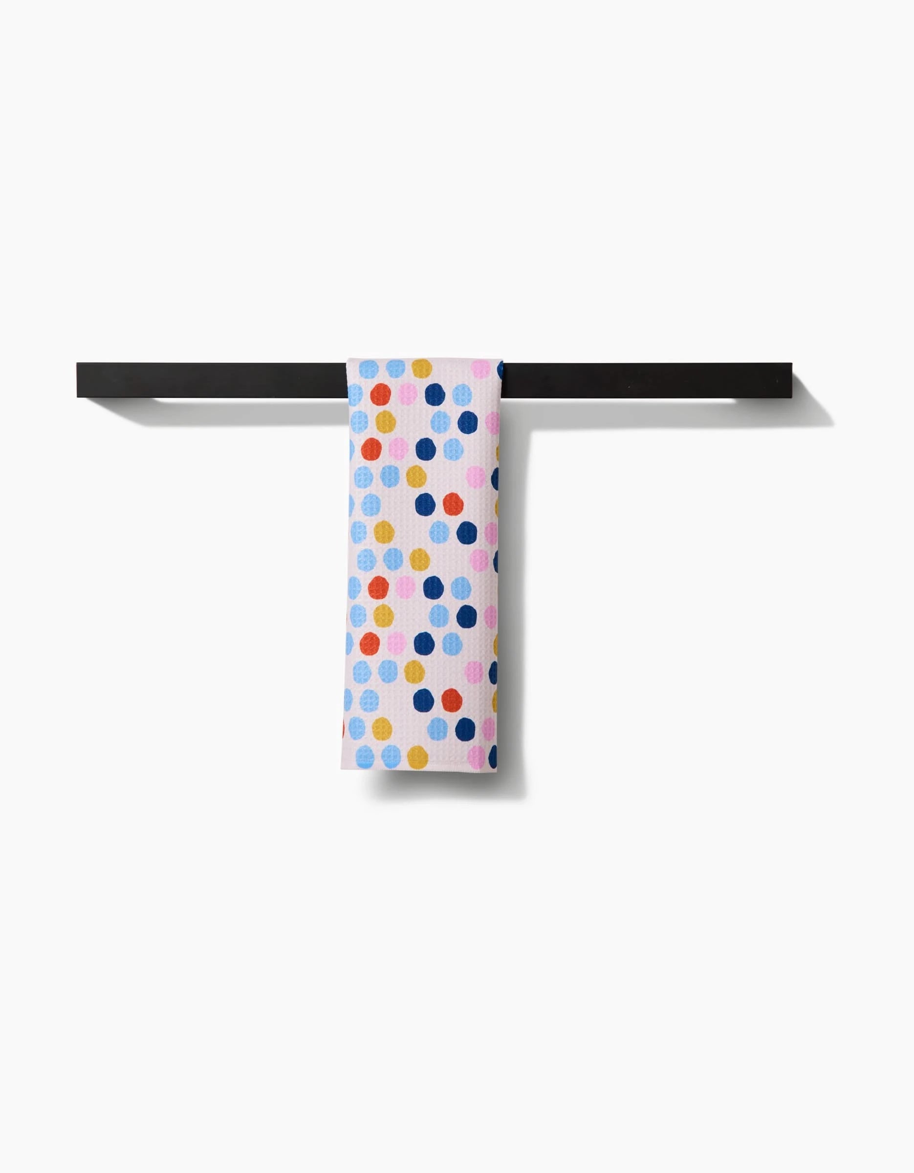 The Carnival bar towel by Lizzie Clark, crafted from recycled materials, features colorful blue, yellow, orange, and pink polka dots on white and hangs neatly over a simple black bar against a white background.