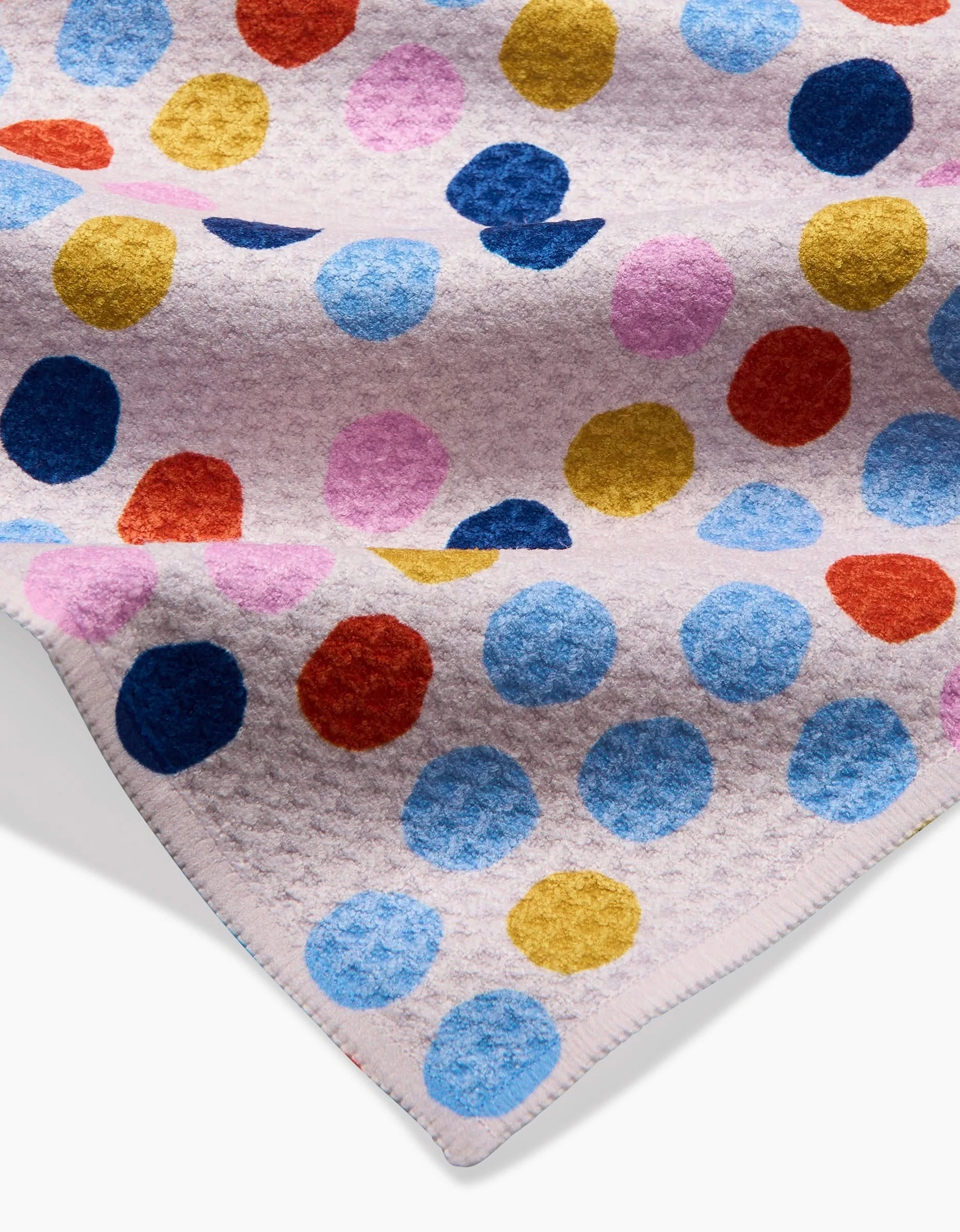 Carnival by Lizzie Clark is a soft, textured blanket made from recycled materials. It features blue, pink, red, and gold polka dots on a light background and has neatly stitched edges for a polished look.