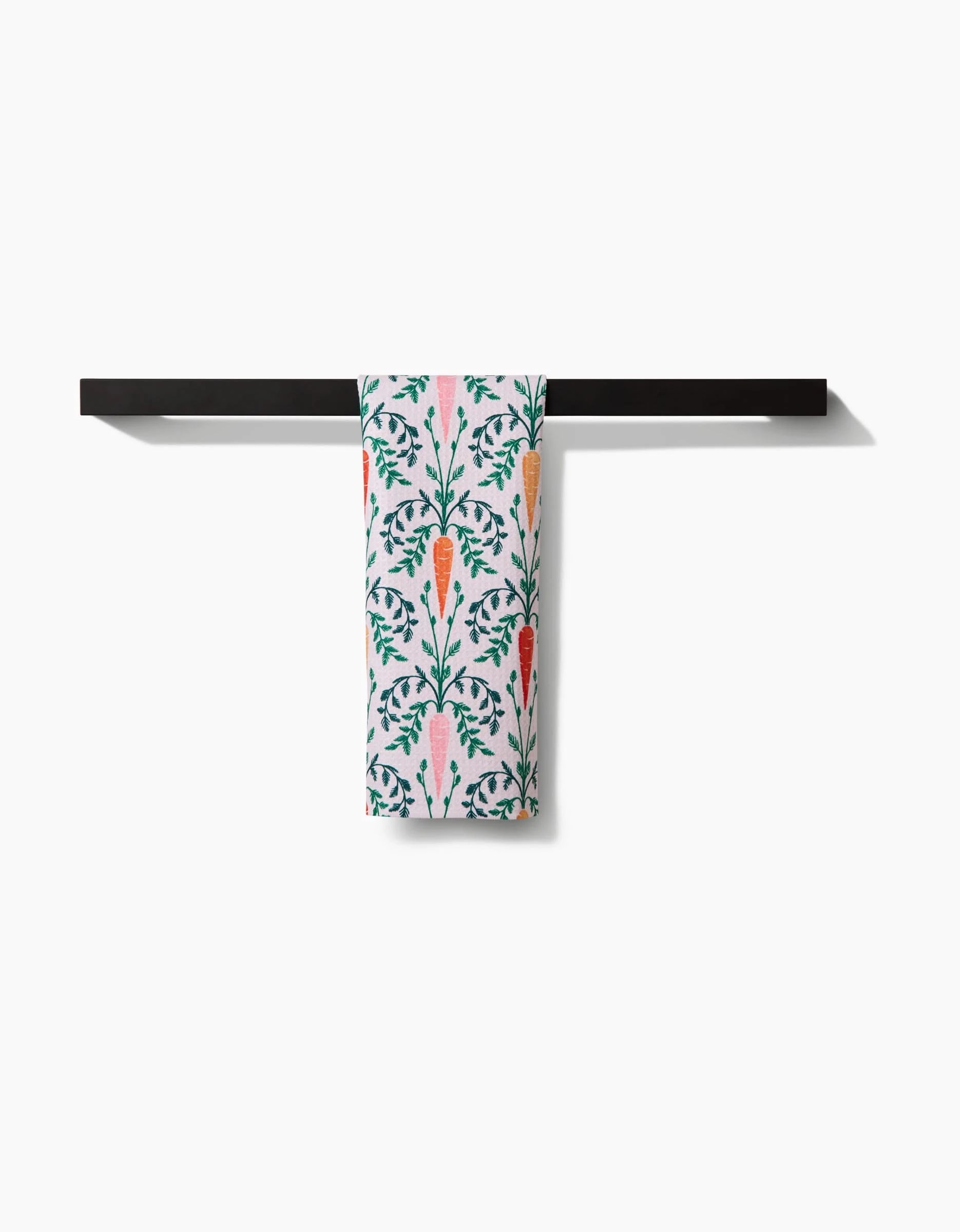The Sarah Daugherty Carrot Damask recycled kitchen towel, featuring vivid orange carrots and green leaves, hangs neatly on a black bar against a plain white background.