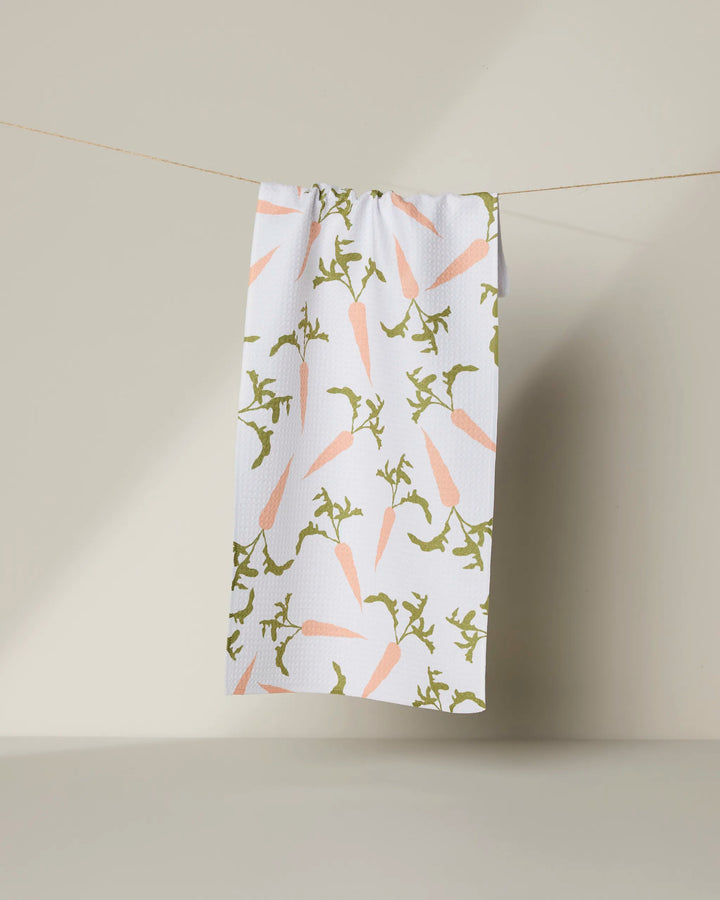 The Geometry Carrot Toss kitchen towel, made from recycled materials, displays light pink carrots and green leaves on white fabric and hangs on a string against a neutral background.