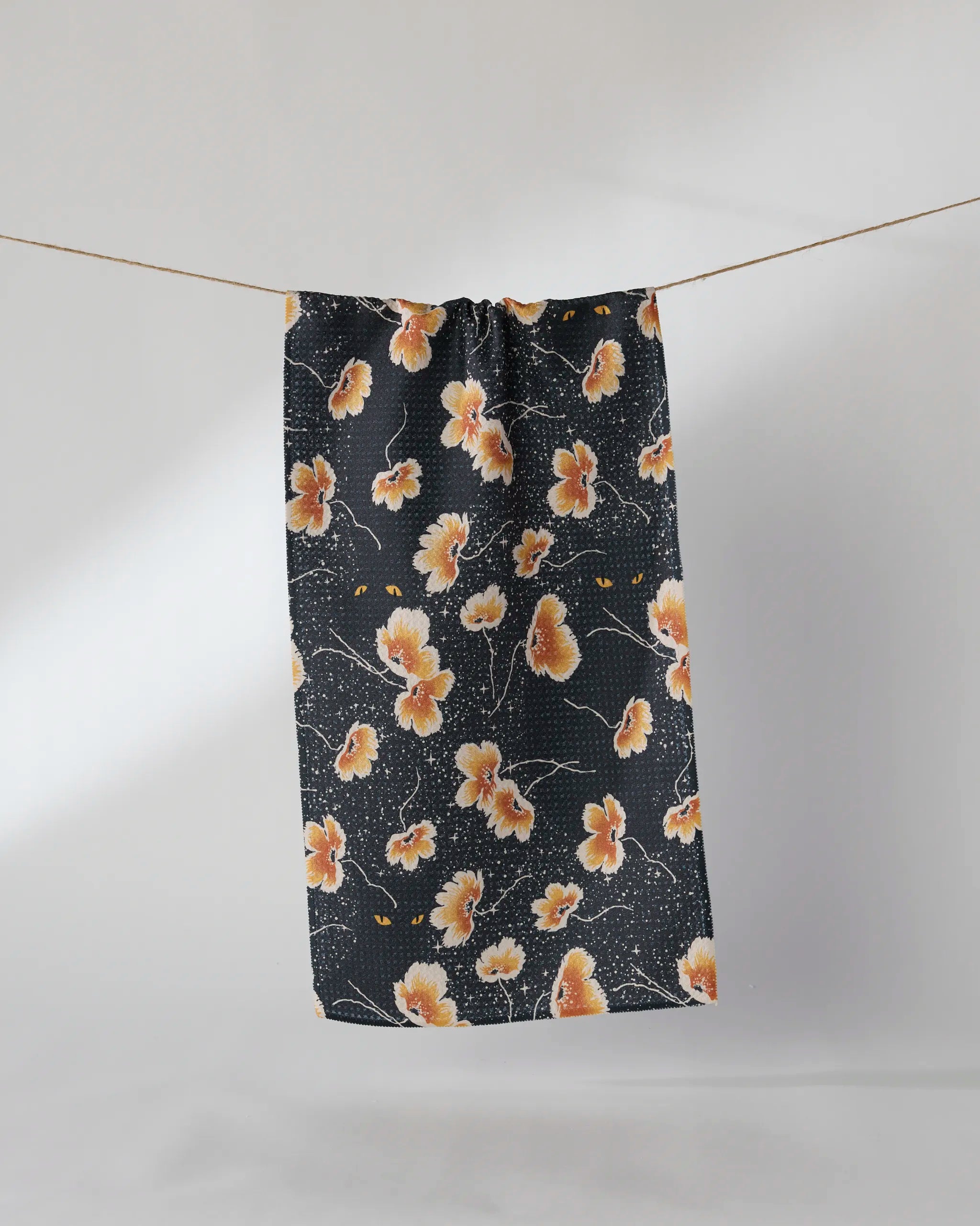 The Geometry Cats Eye recycled kitchen towel, featuring an orange and white floral pattern, hangs on a clothesline against a plain light background.